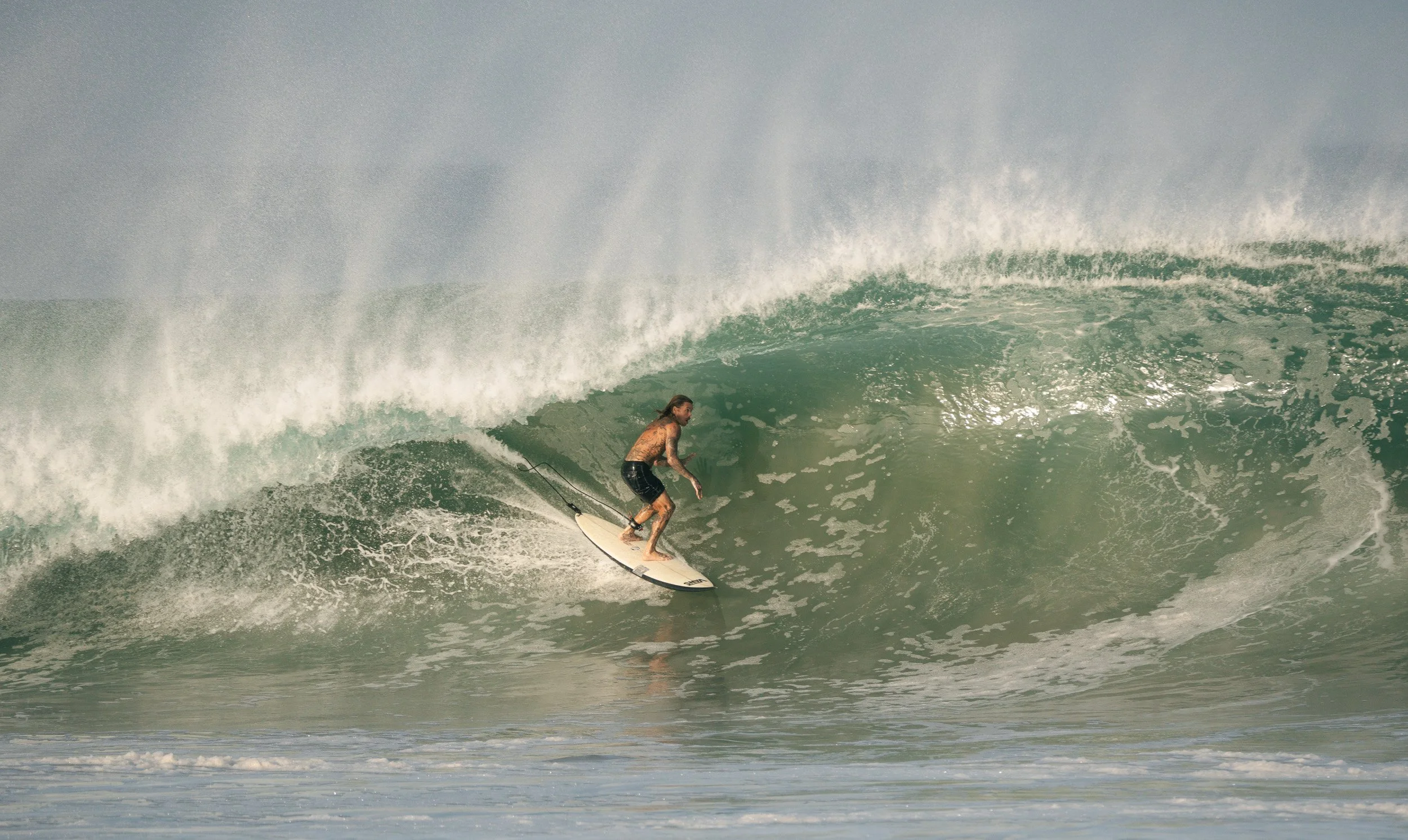 A man with long hair, wearing black shorts, surfing on a large wave in the ocean.