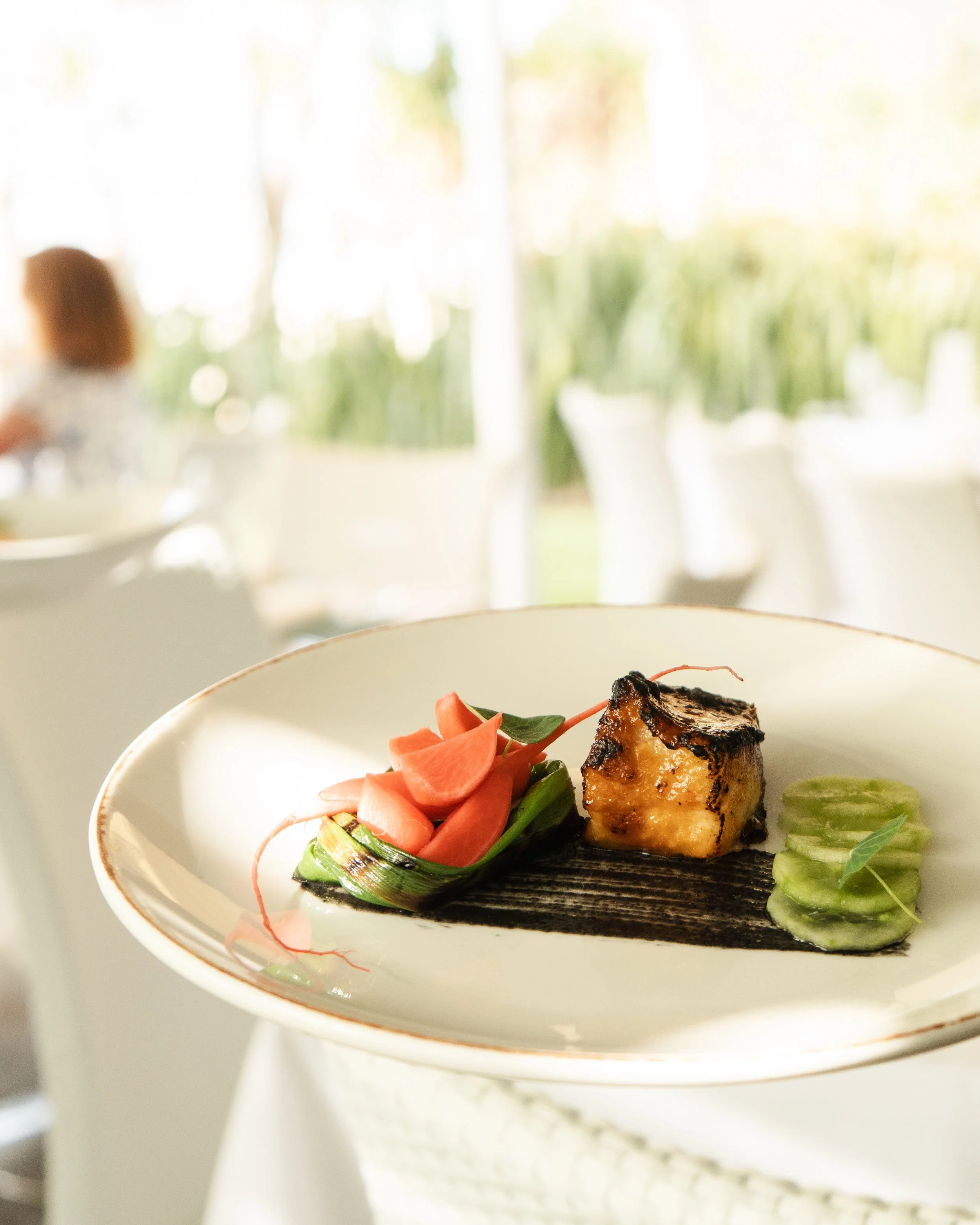 A white plate with a grilled piece of fish, sliced cucumbers, and a vegetable garnish, set against a blurred outdoor dining background.