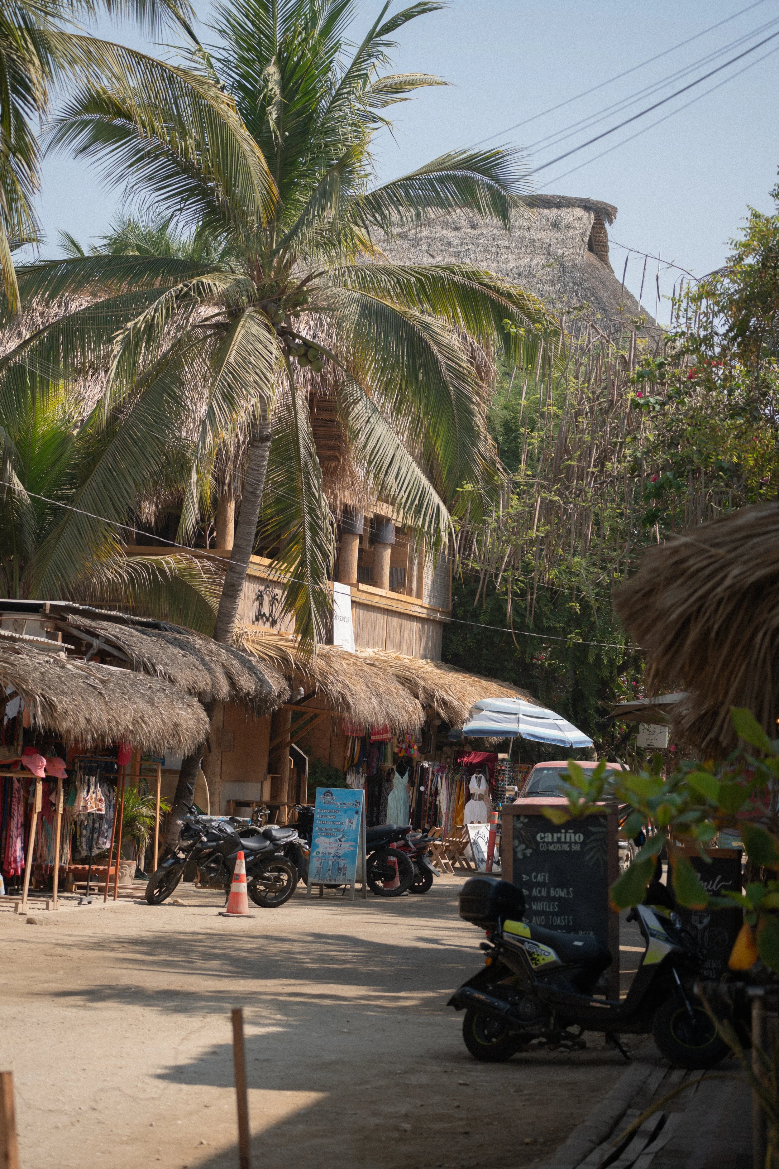 Editorial travel photography of a tropical street with palm trees, thatched-roof shops, parked motorcycles, and beachside cafes in a vibrant coastal area