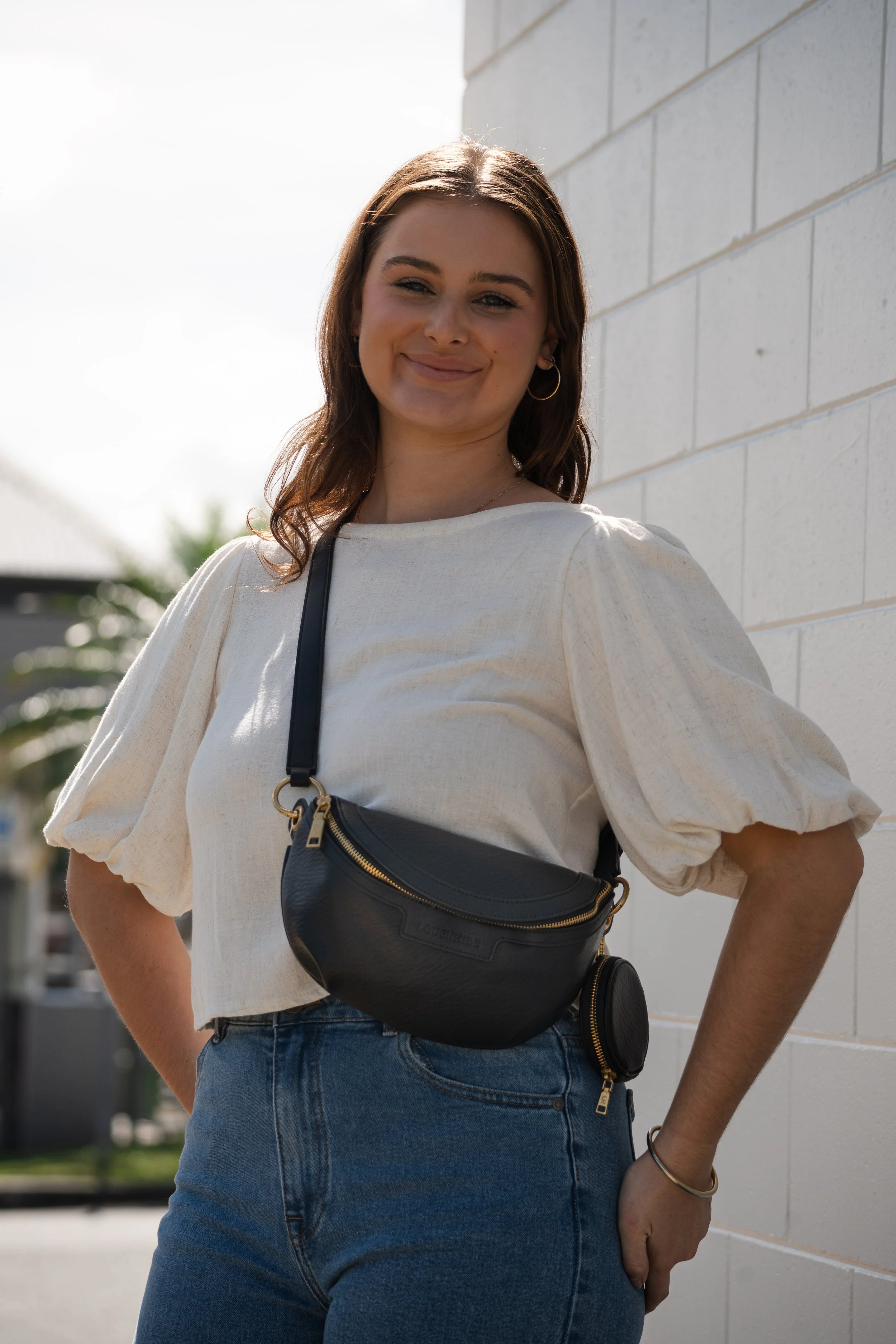A young woman with brown hair and hoop earrings standing outdoors, smiling, wearing a cream-colored blouse, blue jeans, and a black leather crossbody bag with gold zippers, leaning against a white brick wall.