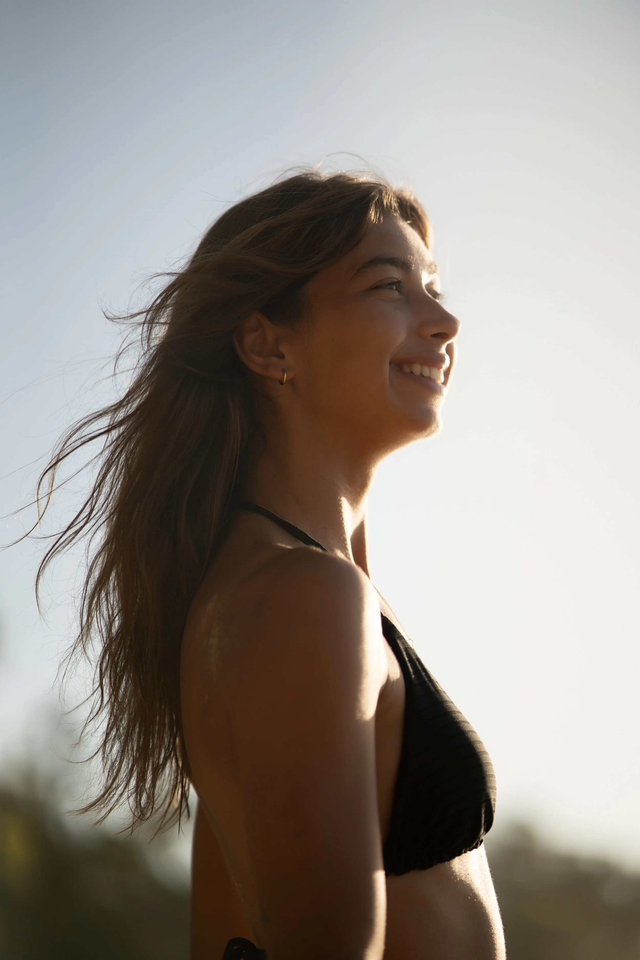 A young woman with long hair smiling, wearing a black bikini top, standing outdoors in sunlight with a clear sky in the background.