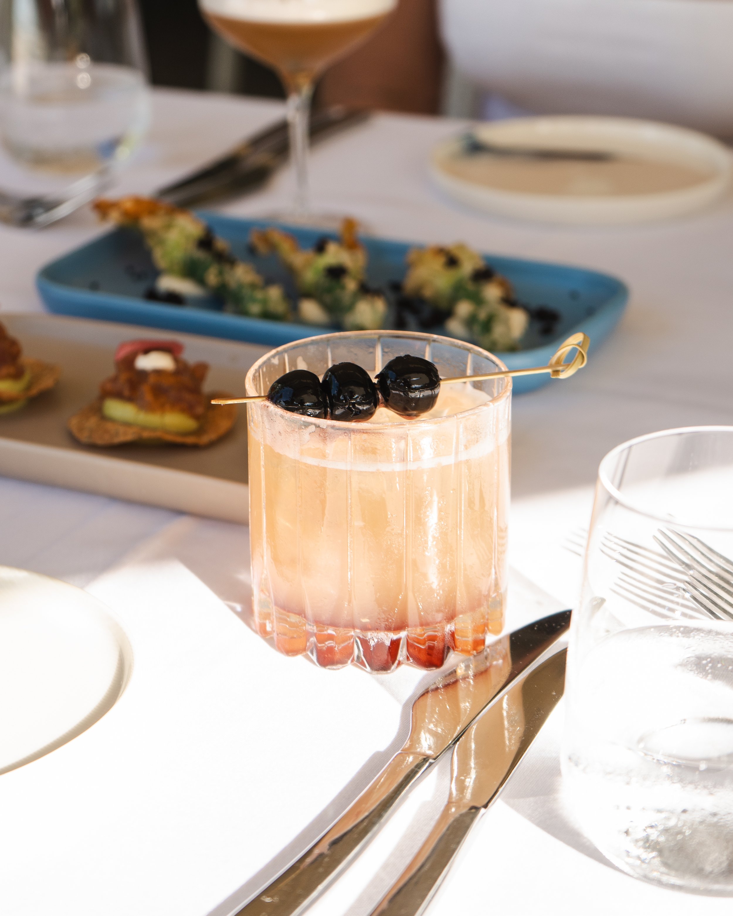 A pink cocktail garnished with three black cherries on a skewer, placed on a table set for dining with utensils, water glass, and plates, with a blurred background of assorted appetizers and sushi.