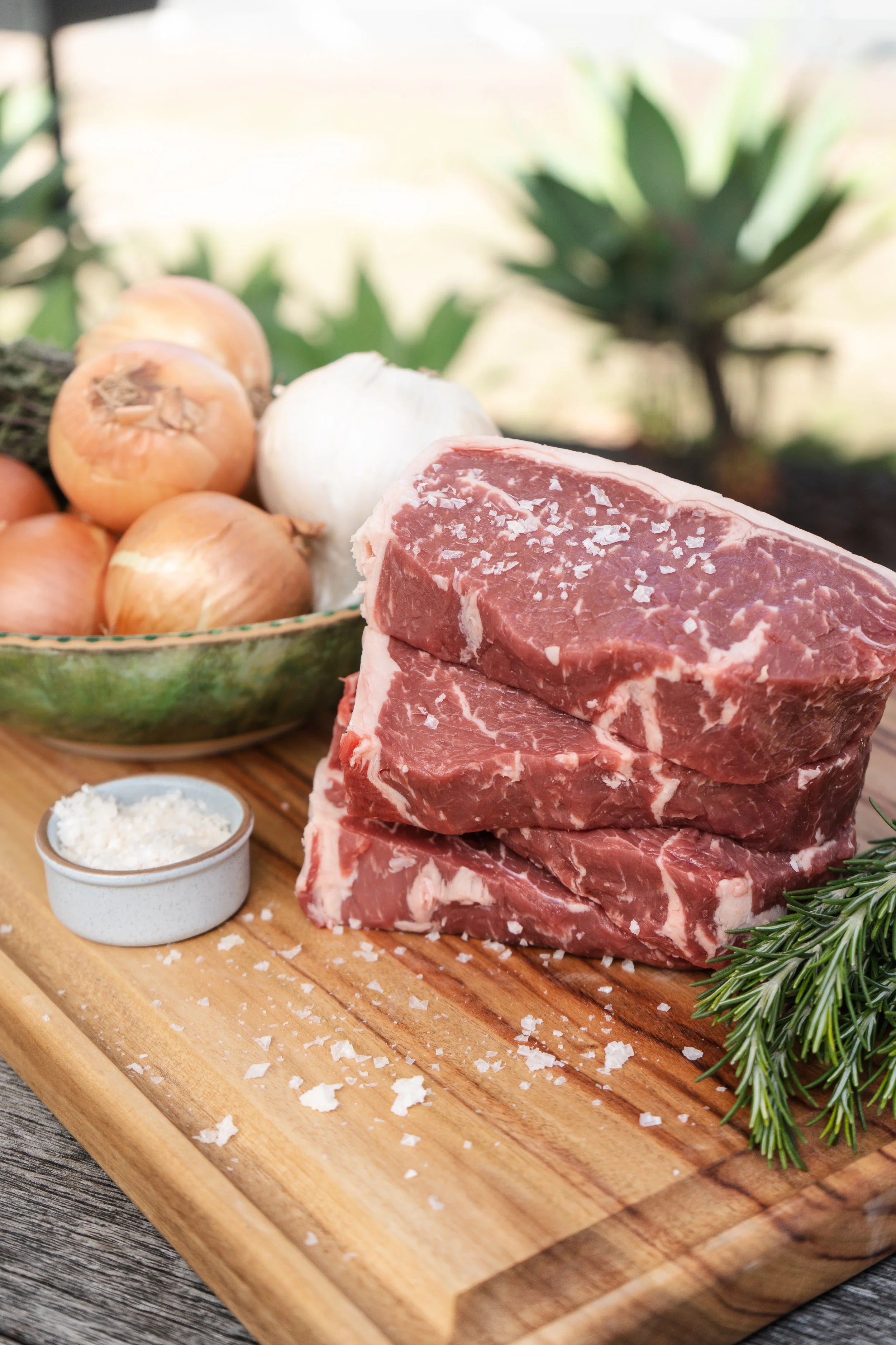 Raw beef steak on a wooden cutting board with onions, garlic, herbs, and seasoning nearby.