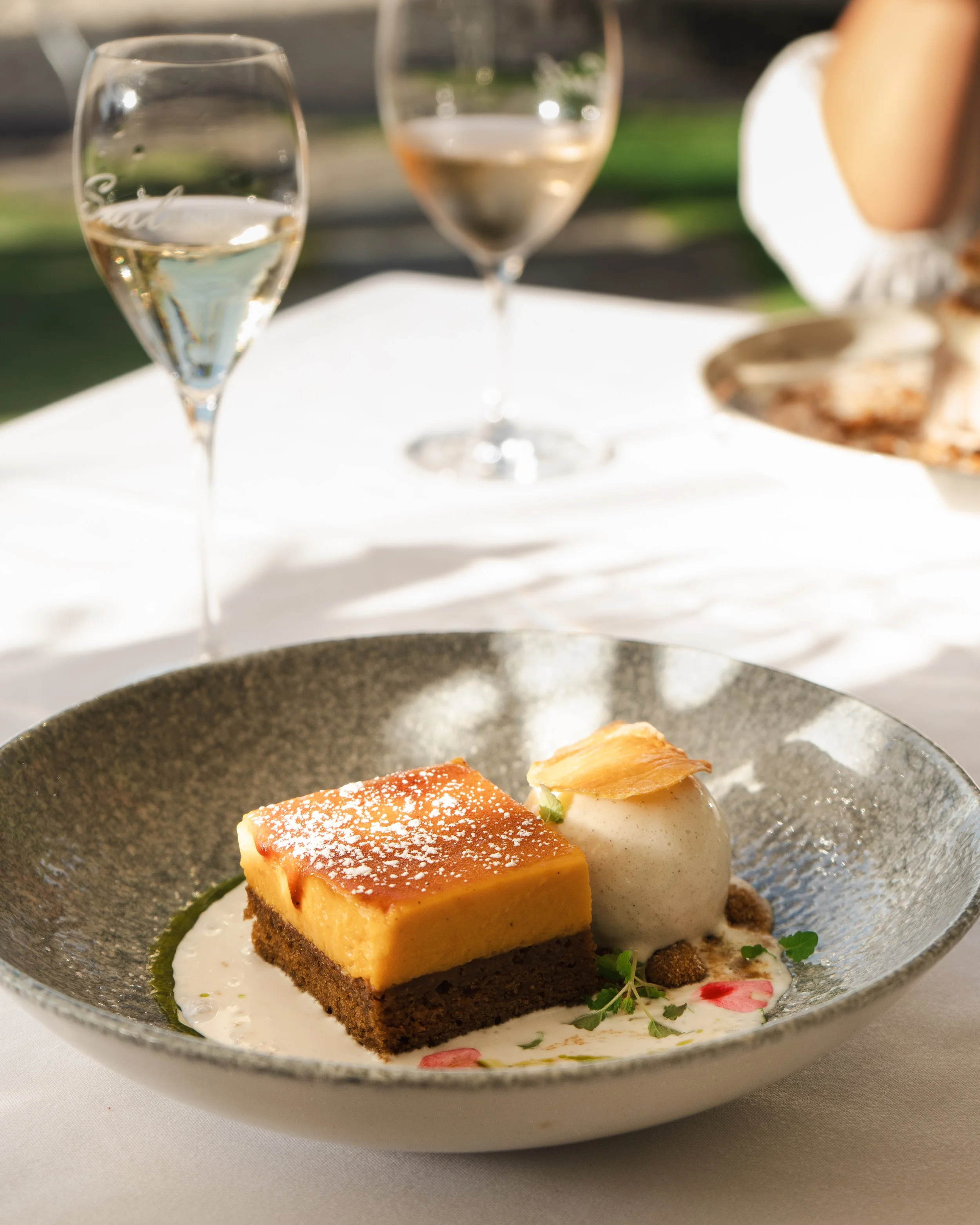 A plated dessert with a layered cake, ice cream, garnishes, and a sauce, on a white table with two glasses of white wine in the background.