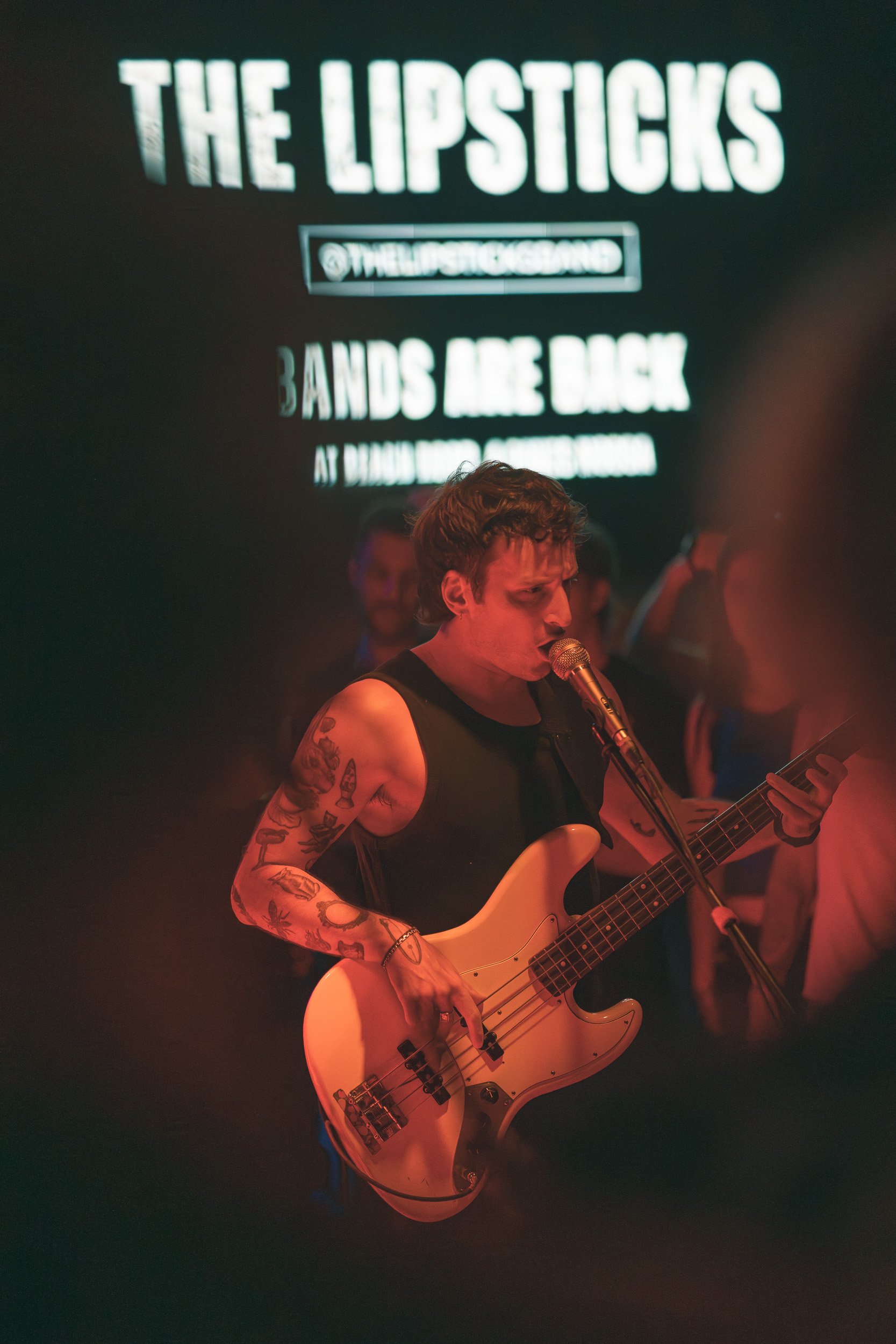 Tattooed musician playing bass guitar and singing into a microphone at a live concert in Noosa, bright ‘The Lipsticks’ sign in background, Sunshine Coast event and music photography