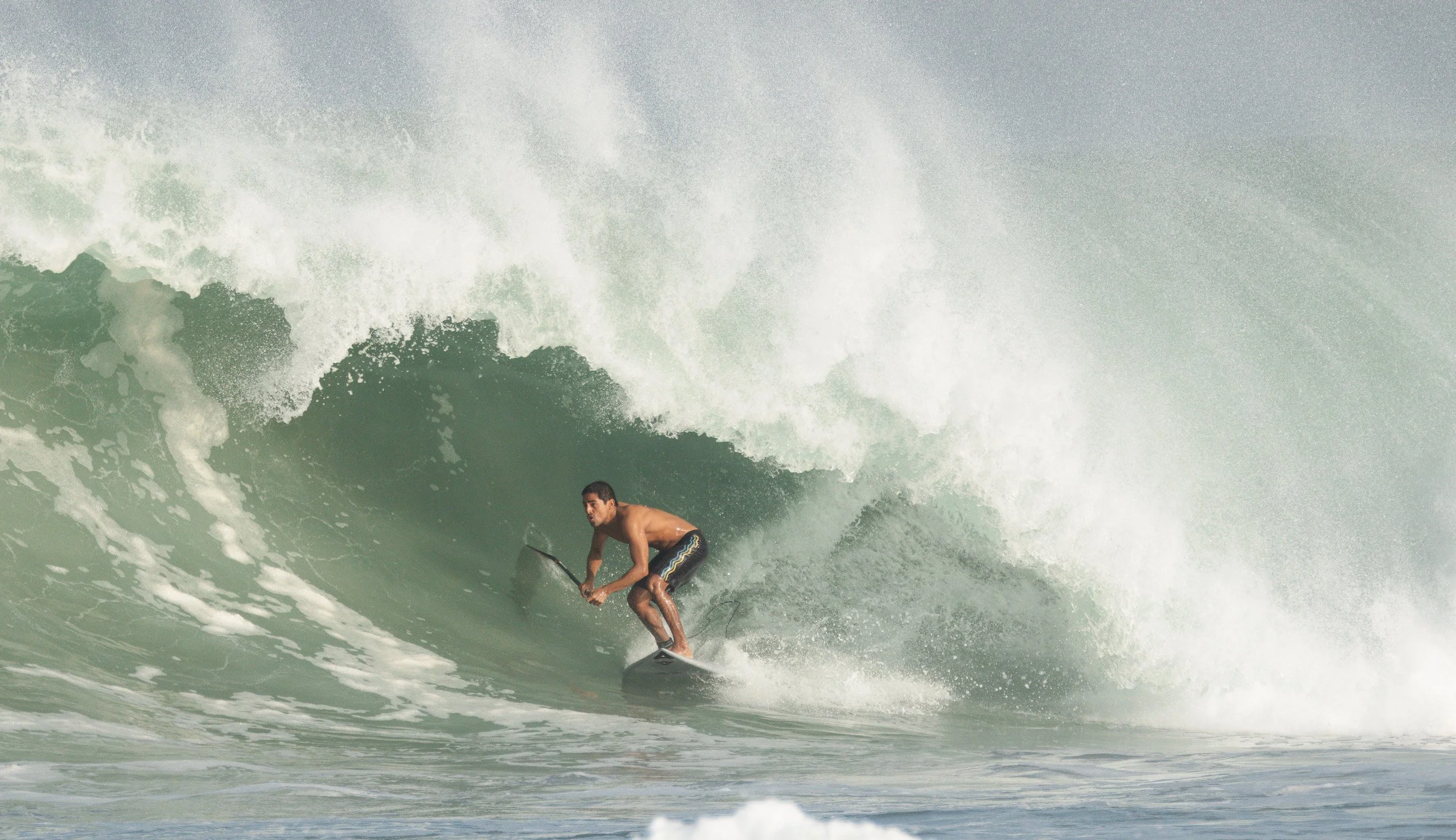 A shirtless man riding a surfboard inside a large breaking wave at the beach.