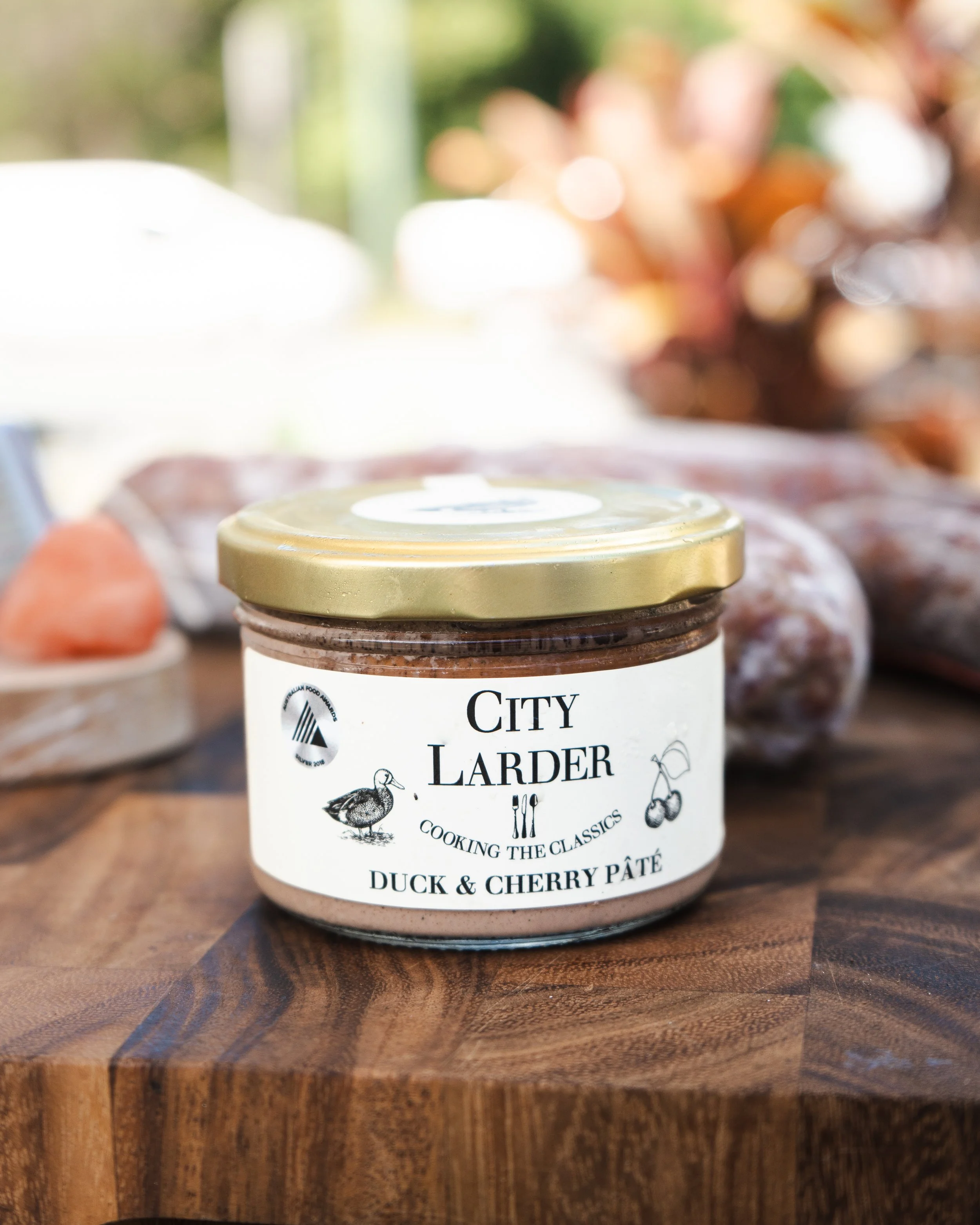 A jar of City Larder duck and cherry pâté on a wooden surface, with blurred background of outdoor setting.