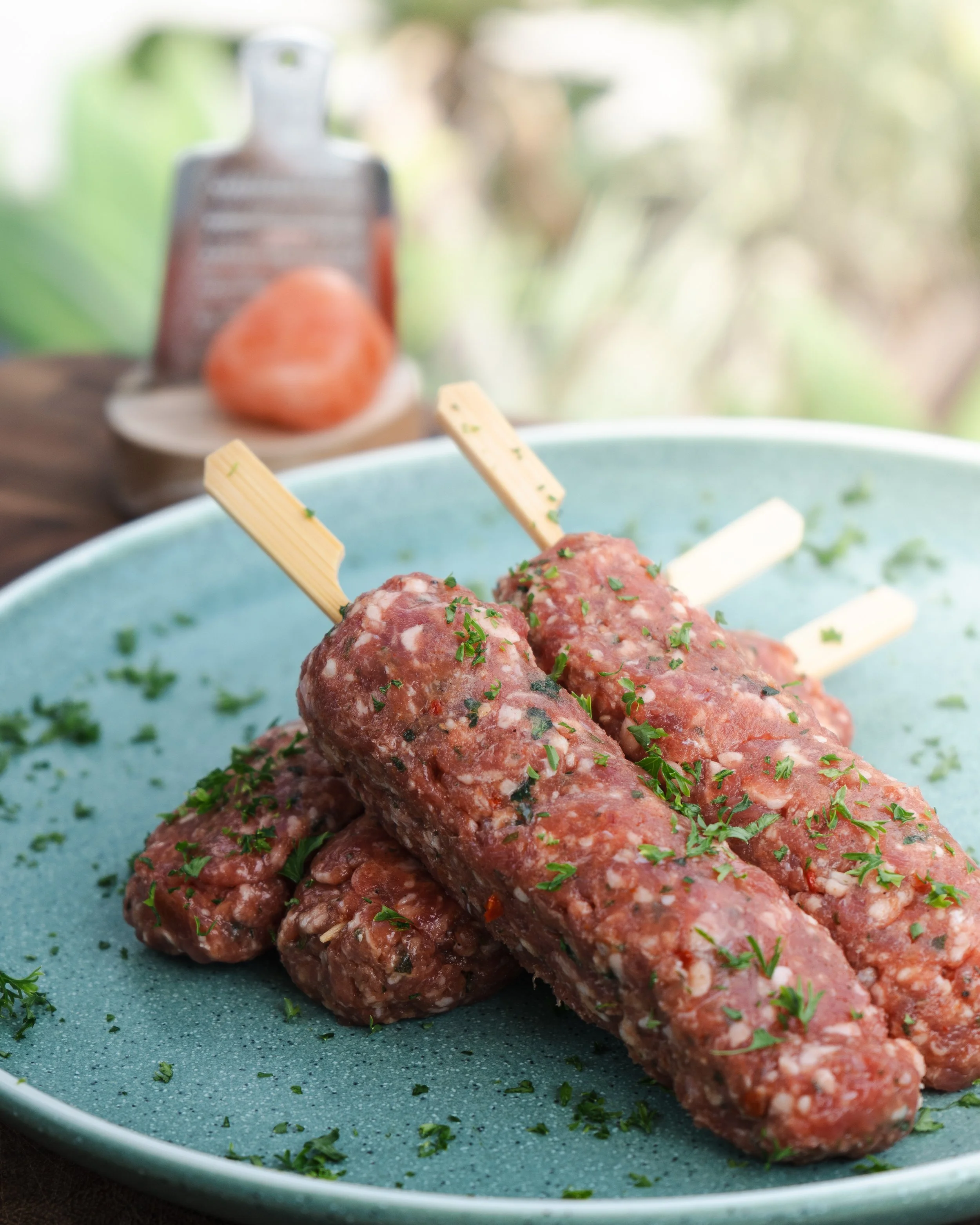 Close-up of raw meatloaf mixture shaped into four logs with wooden sticks inserted, garnished with chopped herbs on a blue plate.