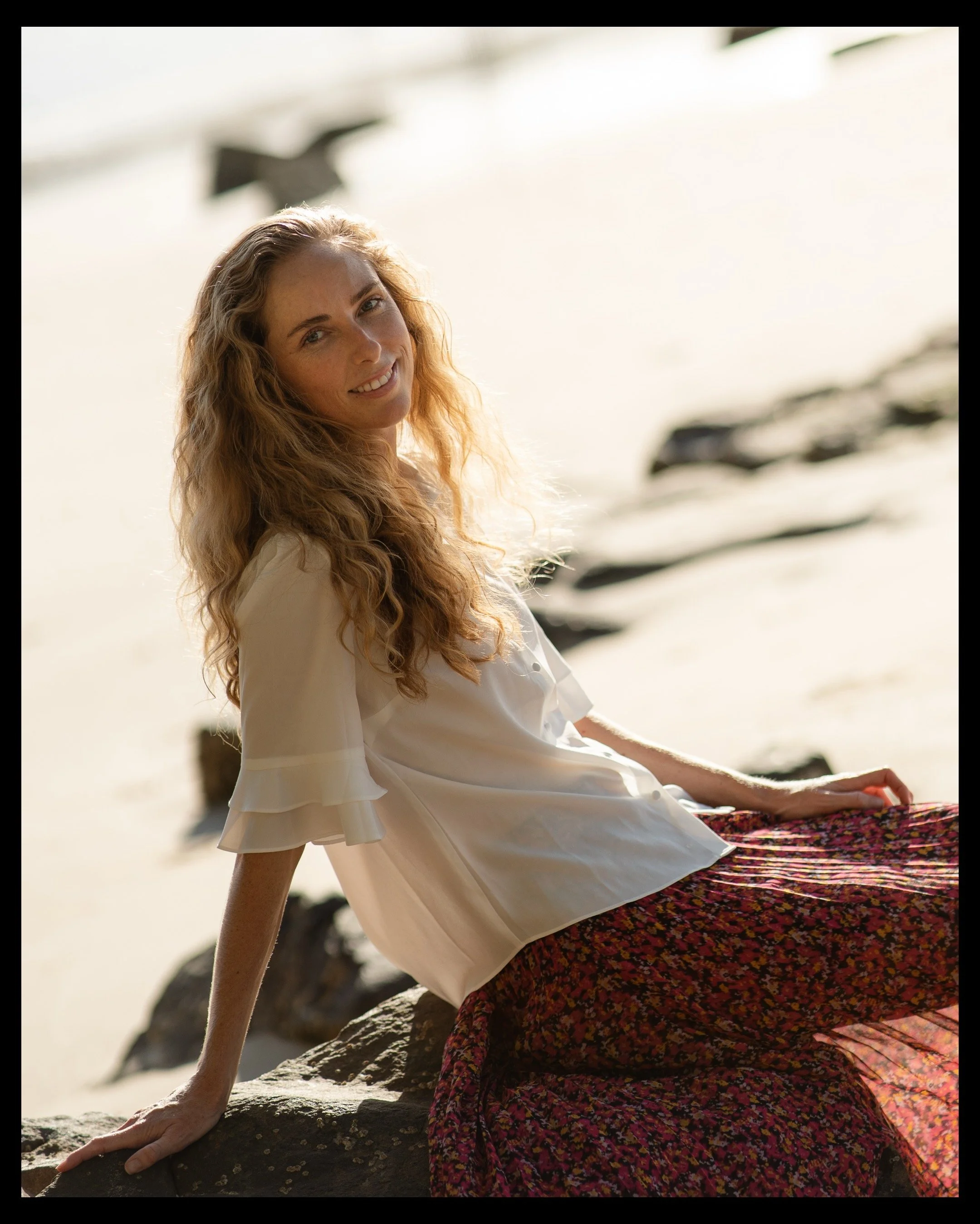 A woman with long curly hair sitting on rocks at the beach, smiling at the camera, wearing a white blouse and a patterned skirt.