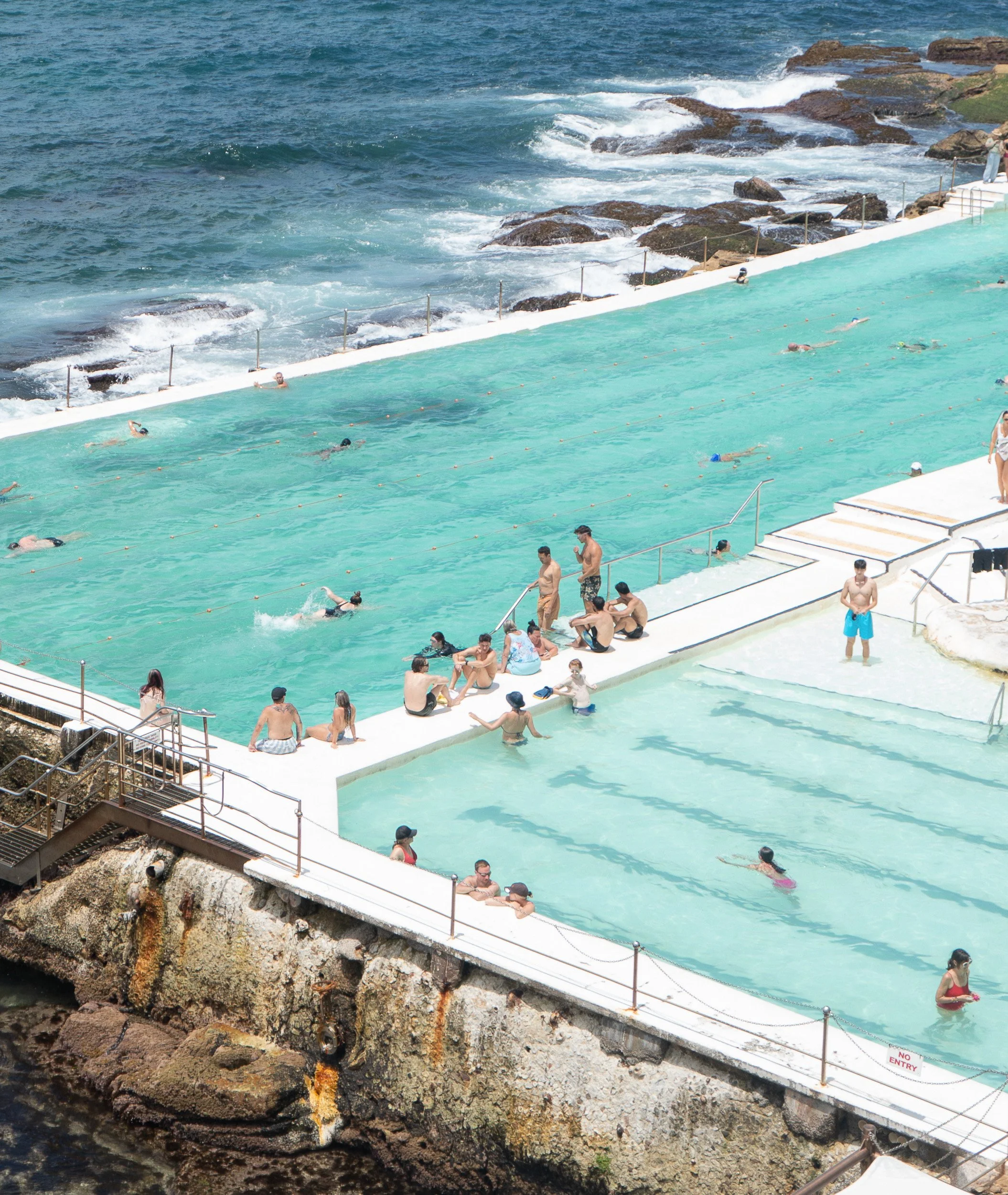 Public swimming pool with people swimming, relaxing, and lounging near the ocean.