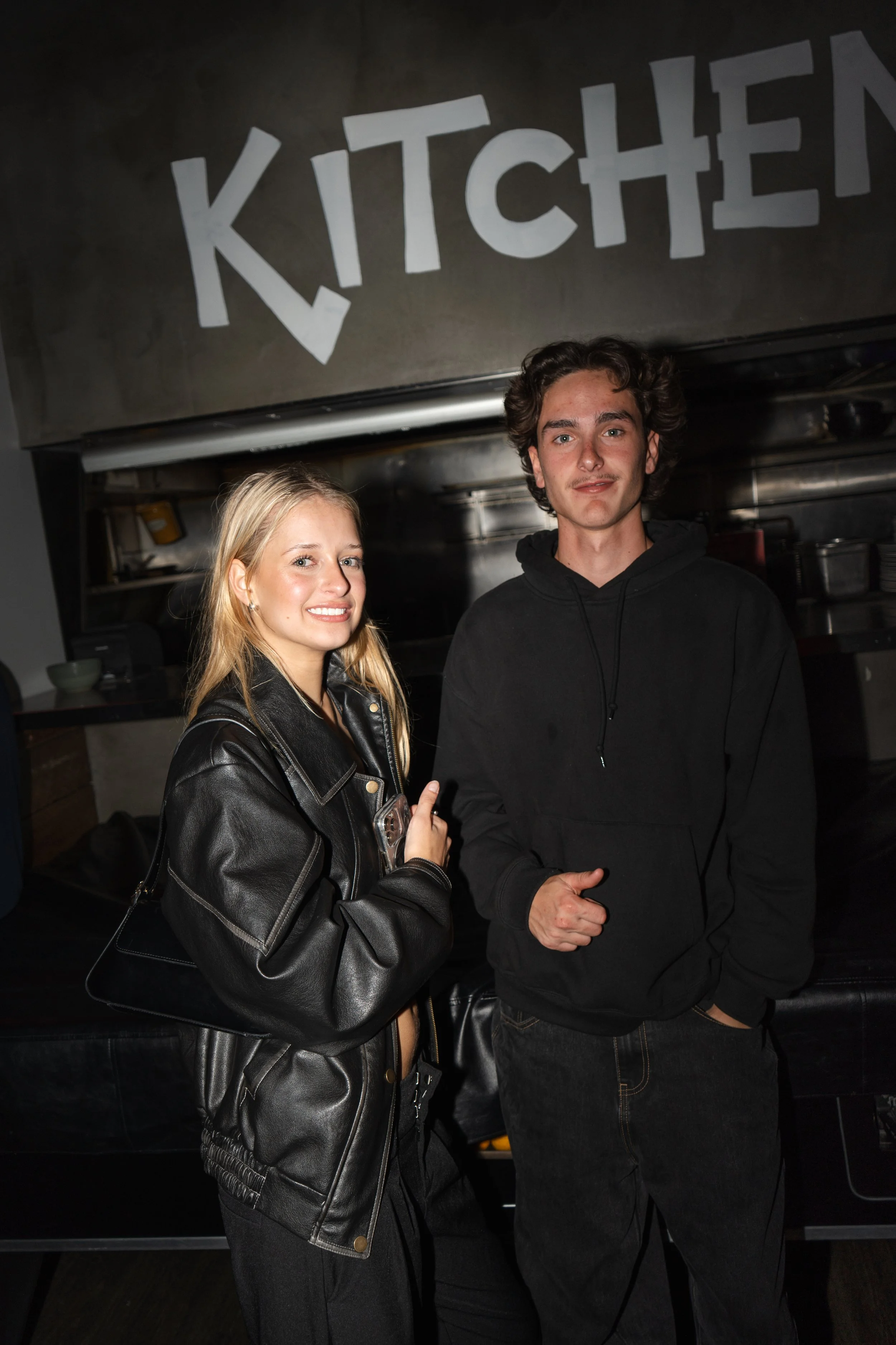 Young woman and man posing together at 4 Pines Noosa with ‘KITCHEN’ sign above in a dark indoor setting, Sunshine Coast event and lifestyle photography