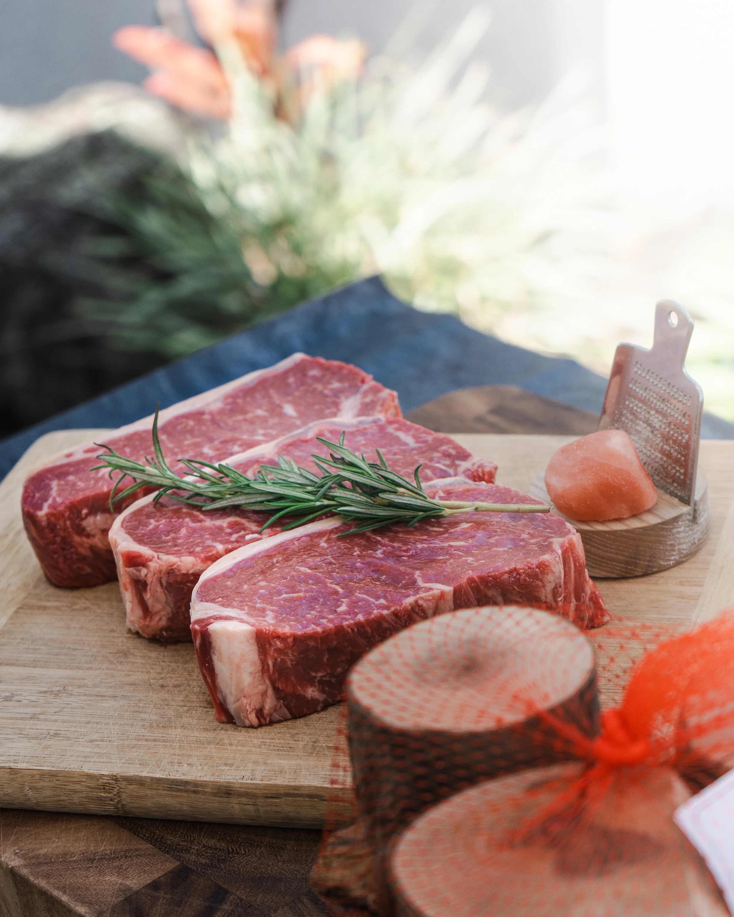 Raw beef steaks with rosemary on a wooden cutting board, salt, and pepper in a shaker, with a salt block and orange netted bag nearby.