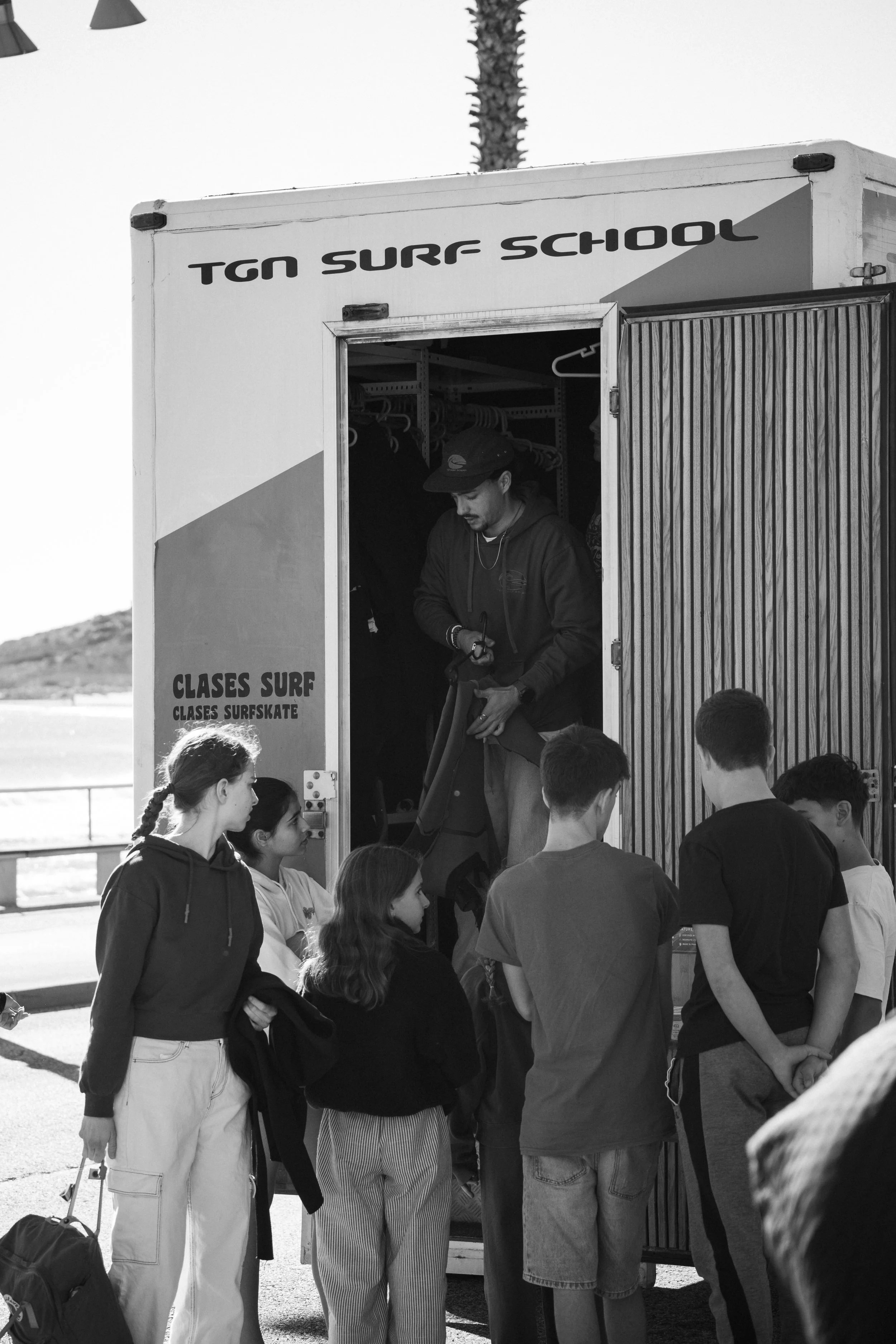 A surf instructor giving lessons to a group of kids at a beach, standing inside a surf school truck labeled "TGN SURF SCHOOL." The kids are wearing casual clothes and holding surfboards or gear.