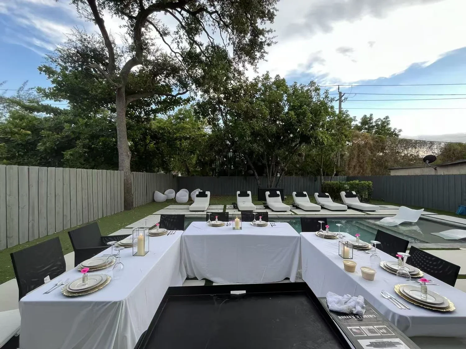 Backyard birthday party setup with a swimming pool, outdoor dining table with white tablecloth, candles, plates, utensils, and glasses, and lounge chairs behind the pool, surrounded by a wooden fence and trees.