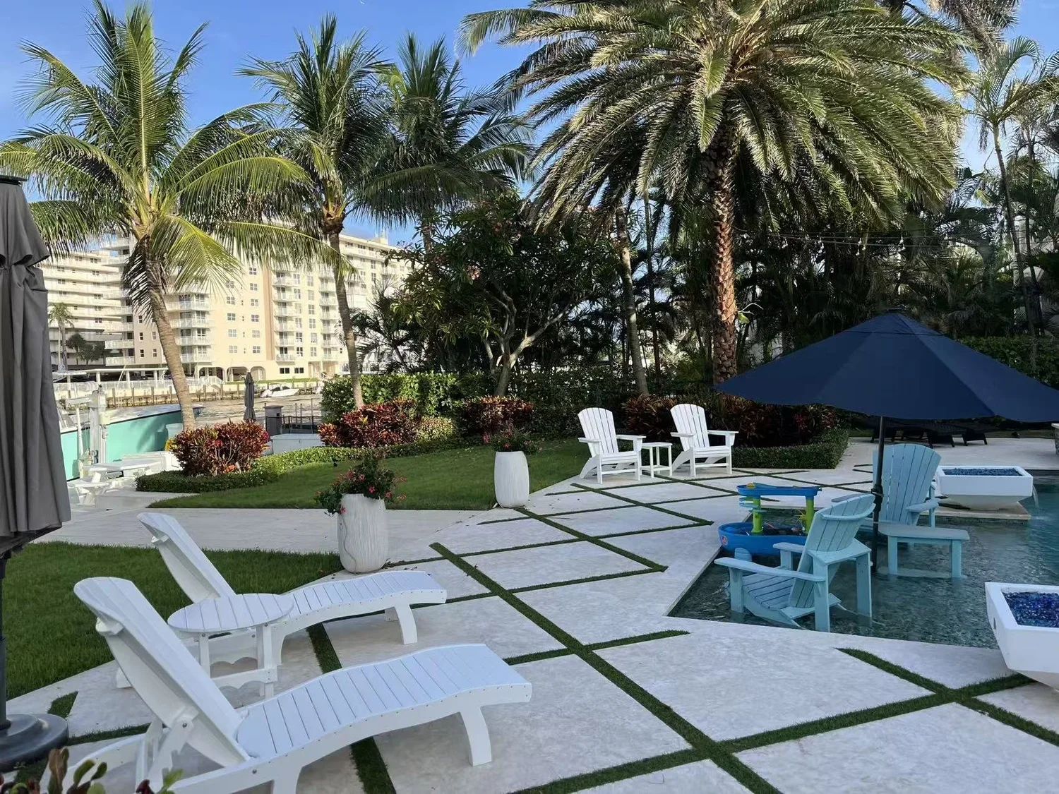 Resort poolside area with white lounge chairs, a blue umbrella, and tropical palm trees in the background under a clear blue sky.