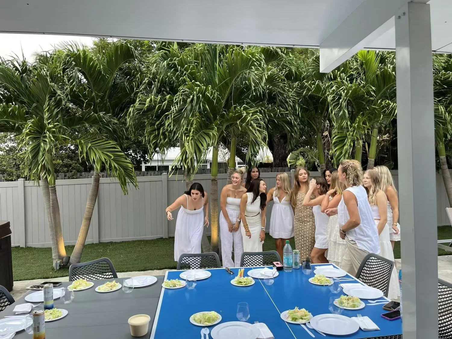 Group of women in white dresses gathered on a backyard patio with palm trees, preparing for a celebration or event.