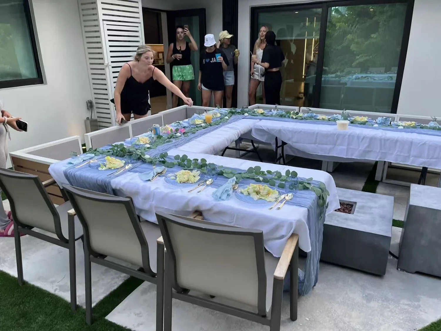 People setting up a long rectangular table decorated with white and blue tablecloths, flower arrangements, and greenery for an outdoor event on a patio.