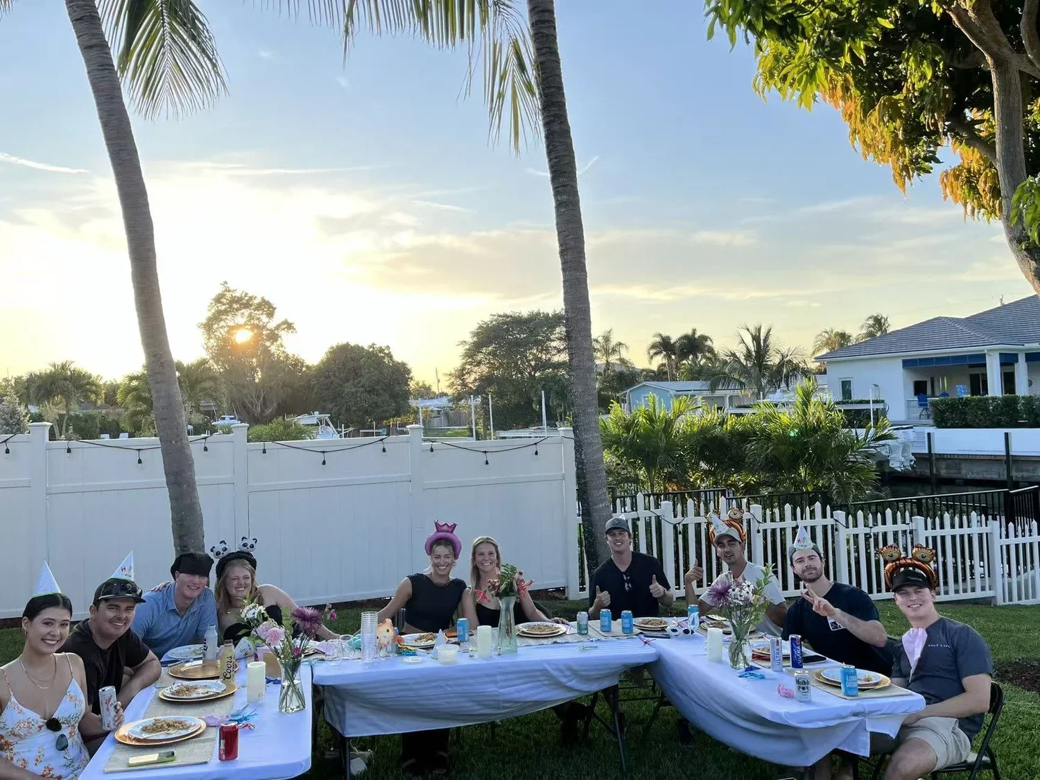 People celebrating a birthday at an outdoor party, sitting at tables with party hats and birthday decorations, in a backyard with palm trees and houses during sunset.