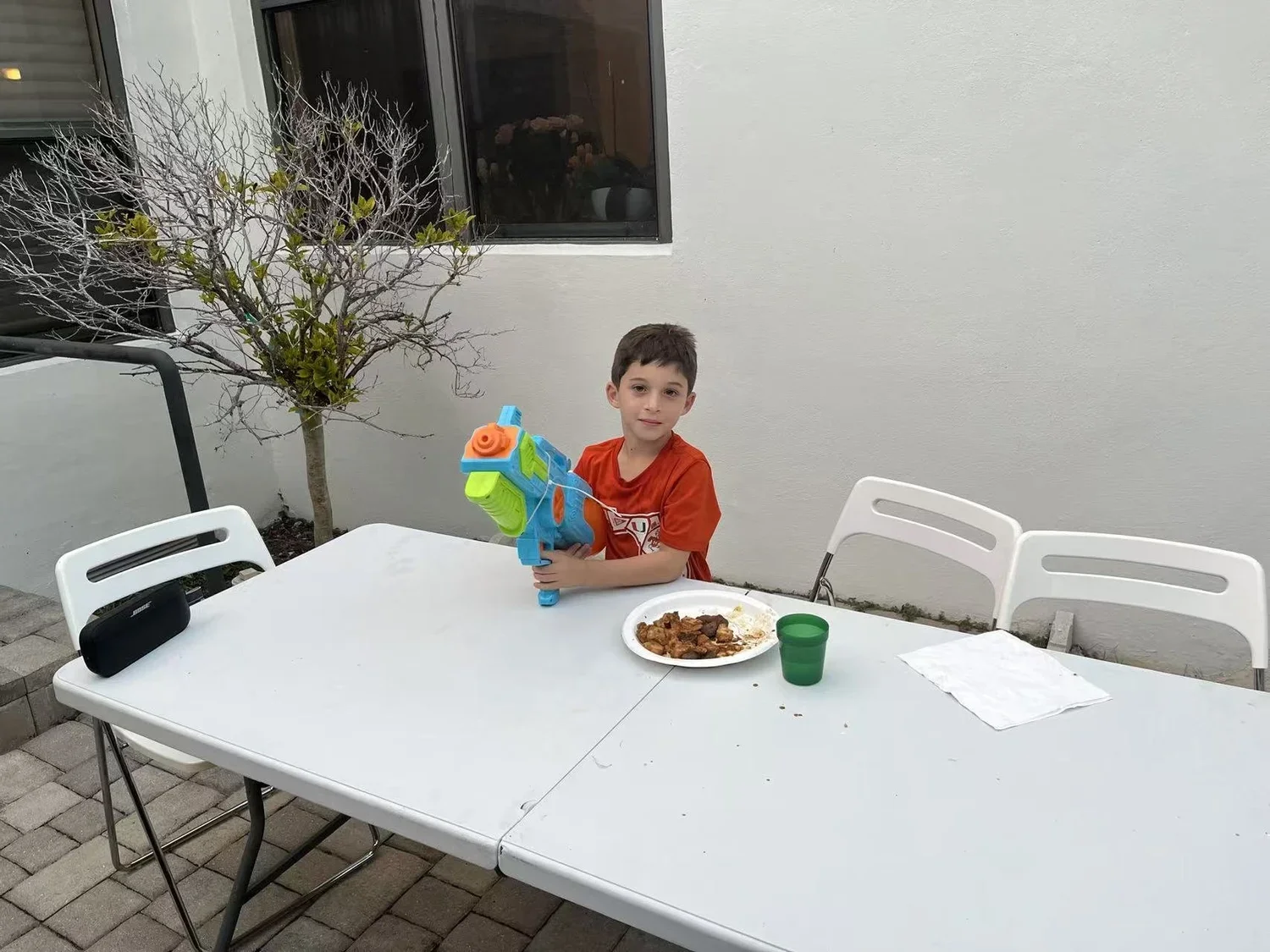 A young boy sitting at a white outdoor table, holding a colorful toy water gun, with a plate of food, a green cup, and a white napkin on the table. There are white chairs and a plant with bare branches in the background.