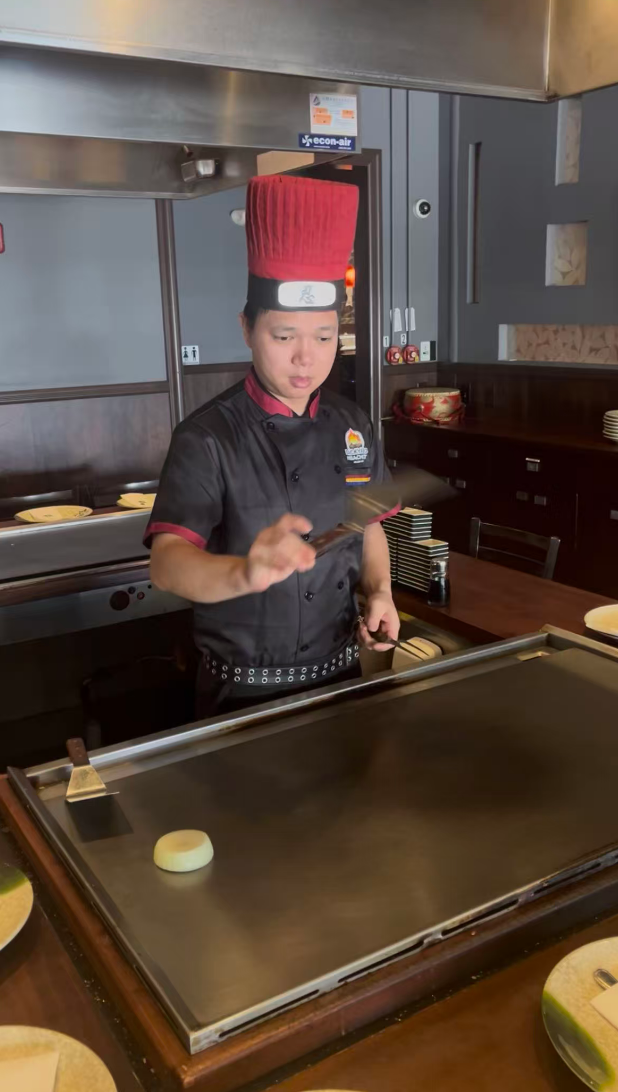 A chef wearing a tall red hat and black uniform prepares food at a grill.