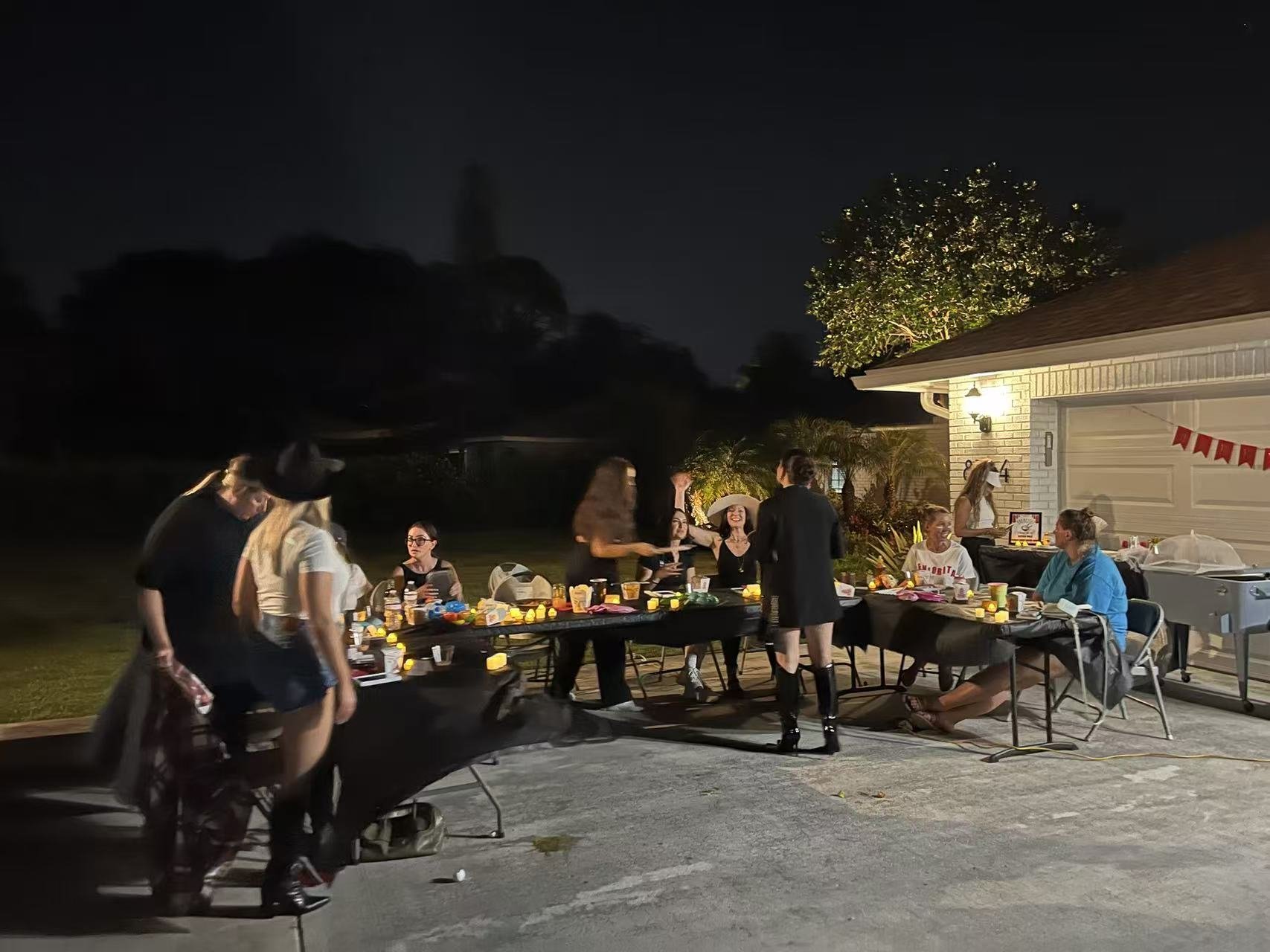 A group of people gathered outside at night for a celebration, sitting and standing around tables with decorations, snacks, and candles, near a house with a garage door and festive banners.