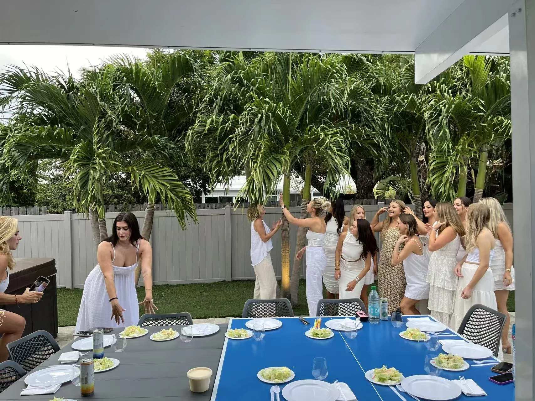 Group of women in white dresses taking photos and smiling in outdoor backyard with palm trees, next to dining table set for a meal.