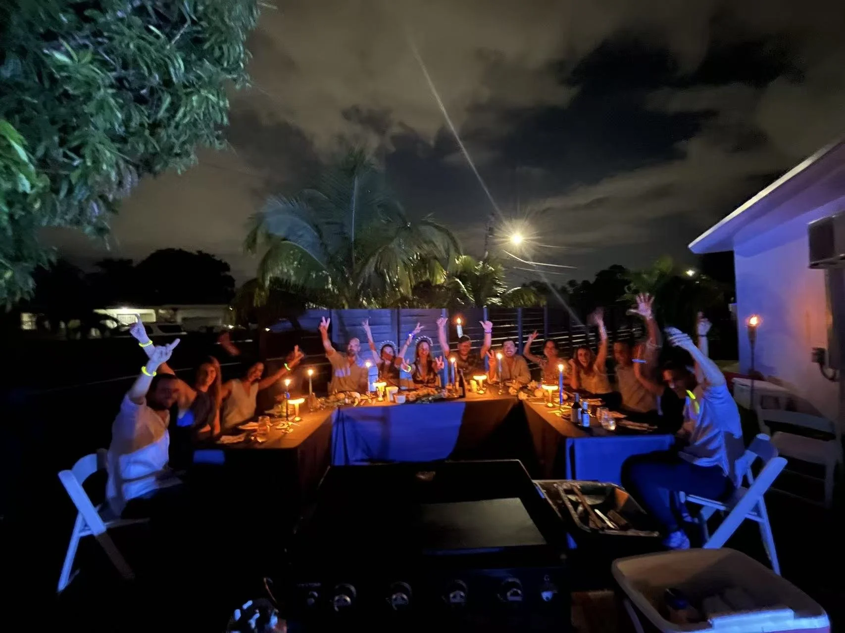 Group of people celebrating at a dinner party outdoors at night, illuminated by candles and string lights, with palm trees and a cloudy sky in the background.