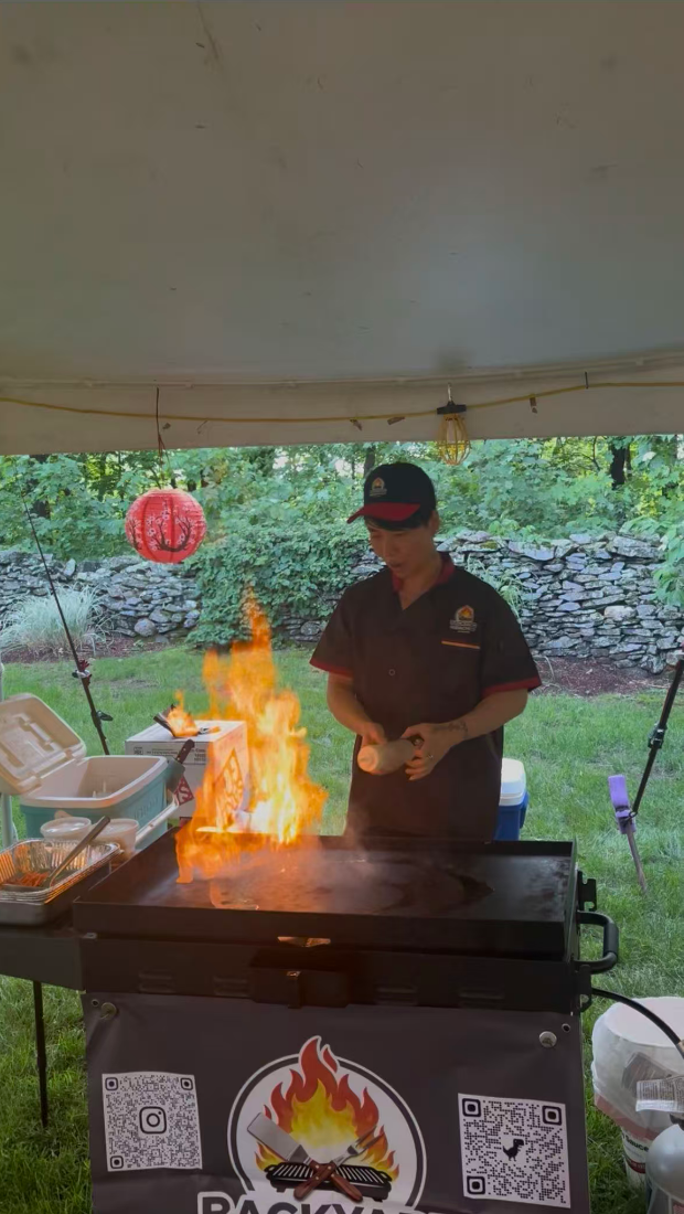 A chef wearing a black and red uniform and cap cooking on a griddle with a fire burning.