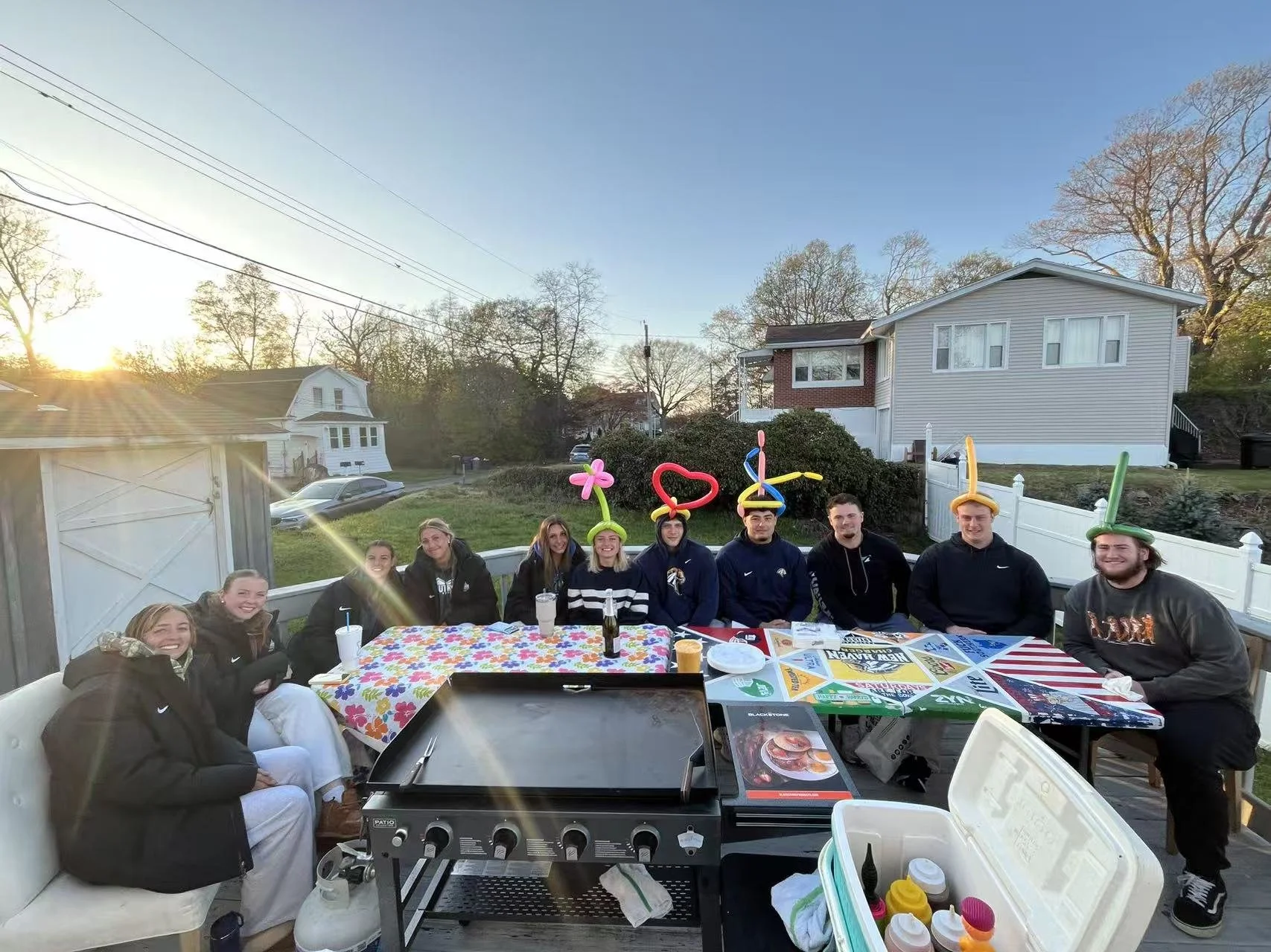 Group of friends gathered around a table outdoors on a porch during sunset, with balloons on their heads, food, drinks, and a grill in front of them, in a neighborhood with houses and trees.