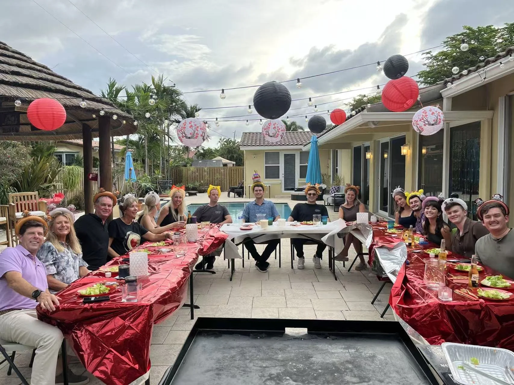 A diverse group of people at a birthday party with animal ear headbands, gathered around a decorated table outdoors in a backyard with a pool and string lights, during daytime.