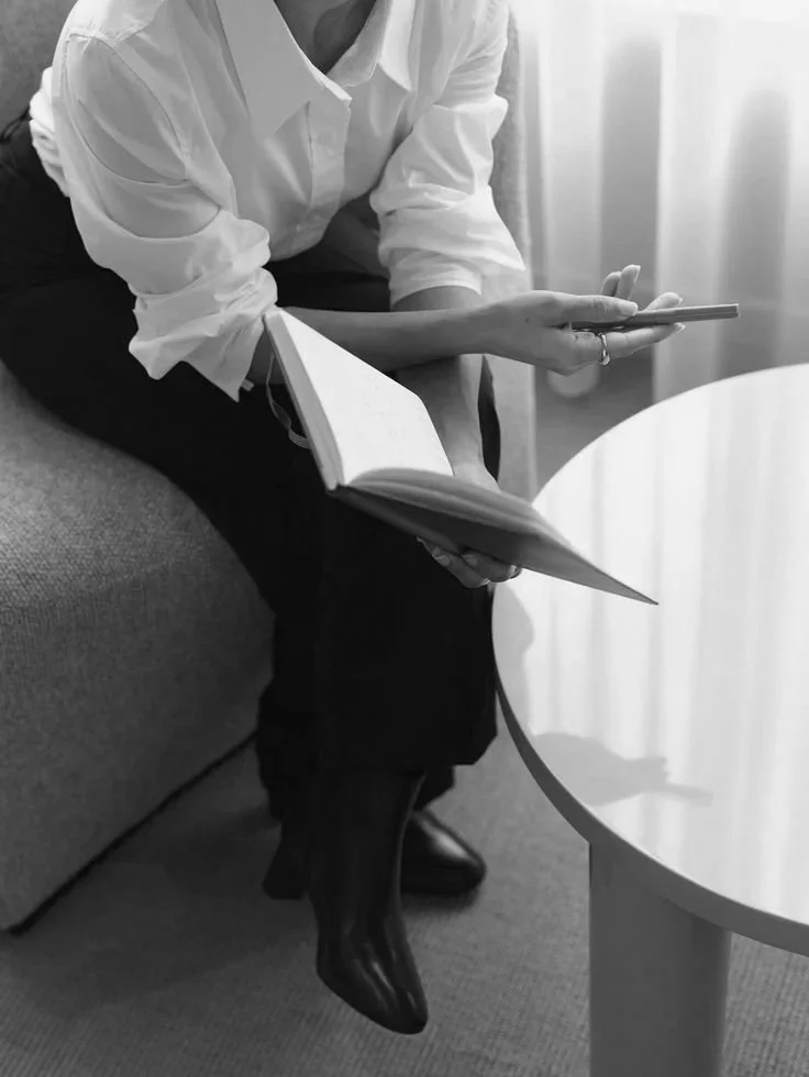 A person sitting on a sofa, holding a notebook in one hand and a smartphone in the other, with a round table nearby, in black and white.