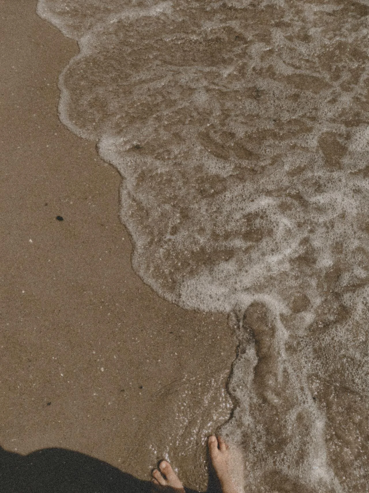 Feet standing on wet sand at the edge of the shoreline, with small waves approaching.