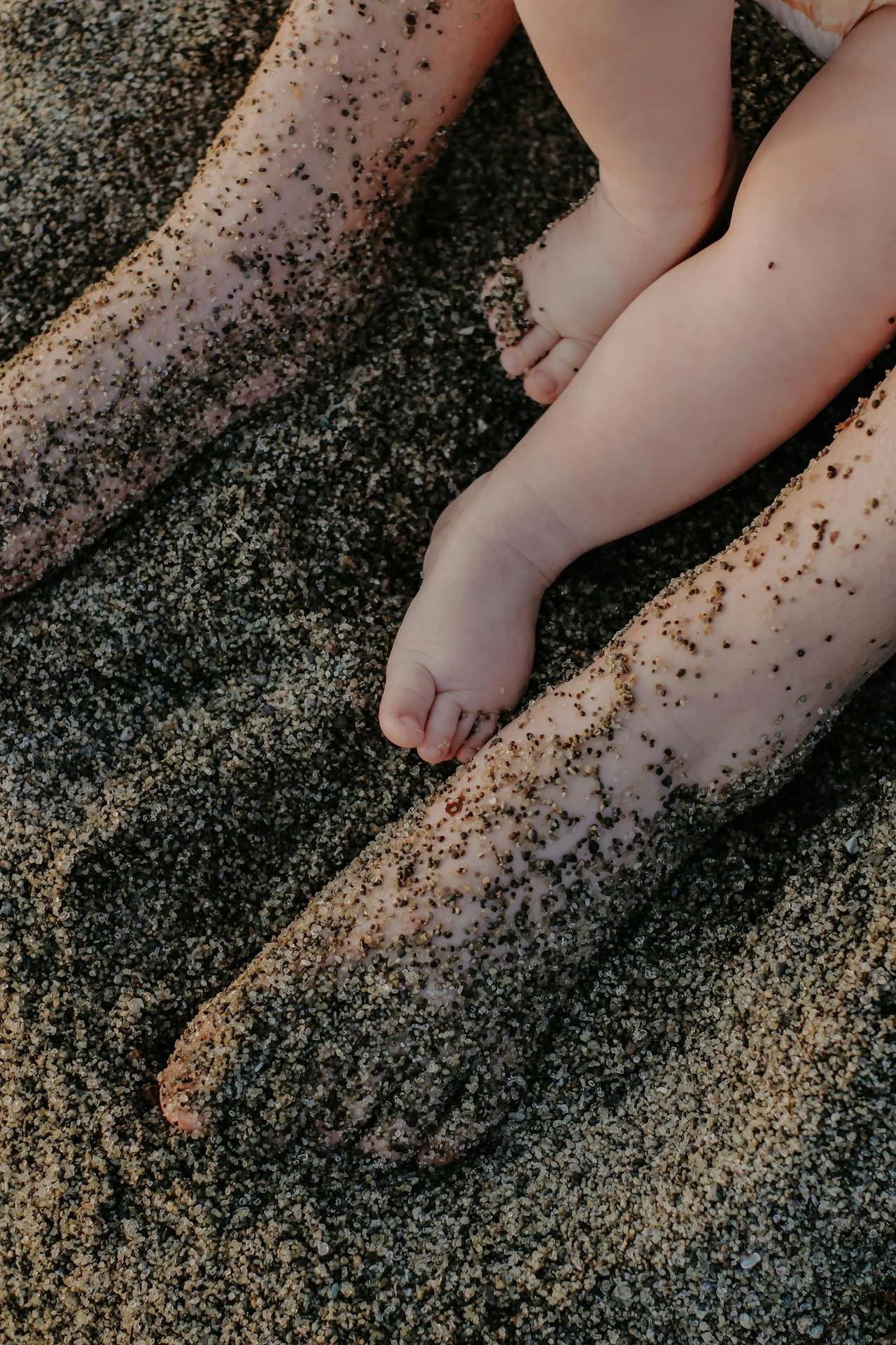 Close-up of a child's hand resting on dad's leg covered in sand, with beach sand and pebbles surrounding them.