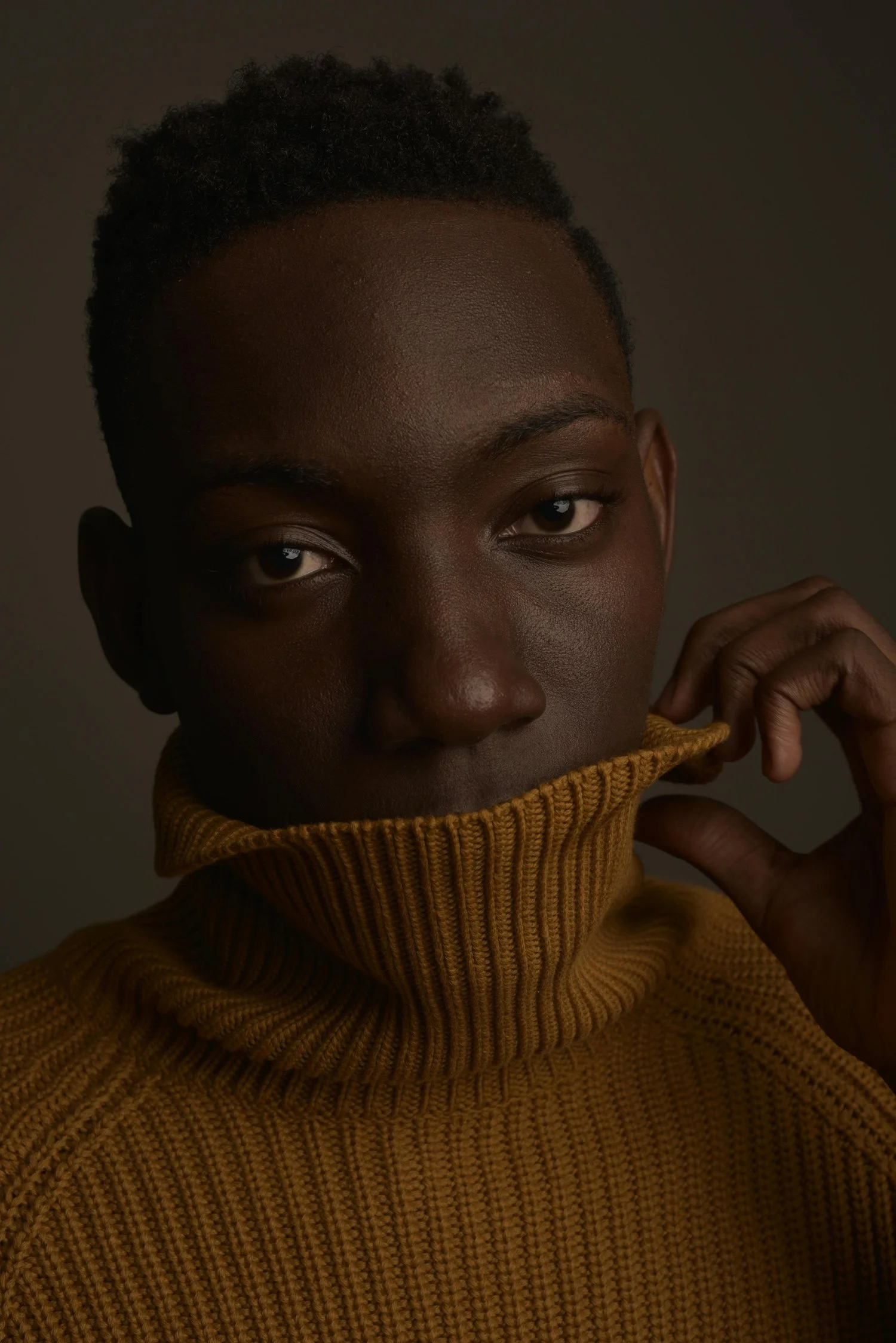 Close-up portrait of a young Black person with short, natural hair, wearing a yellow turtleneck sweater, partially covering the face with the collar, looking directly at the camera.