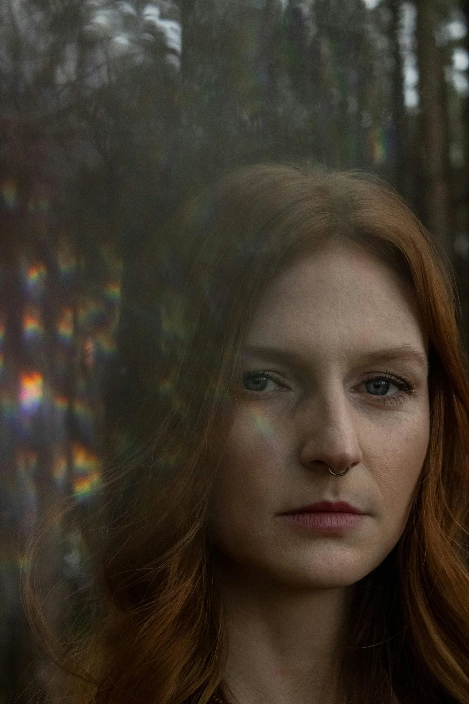 Close-up of a woman with red hair, blue eyes, and a nose ring, standing outdoors with trees in the background. There are rainbow light reflections on the left side of the image.