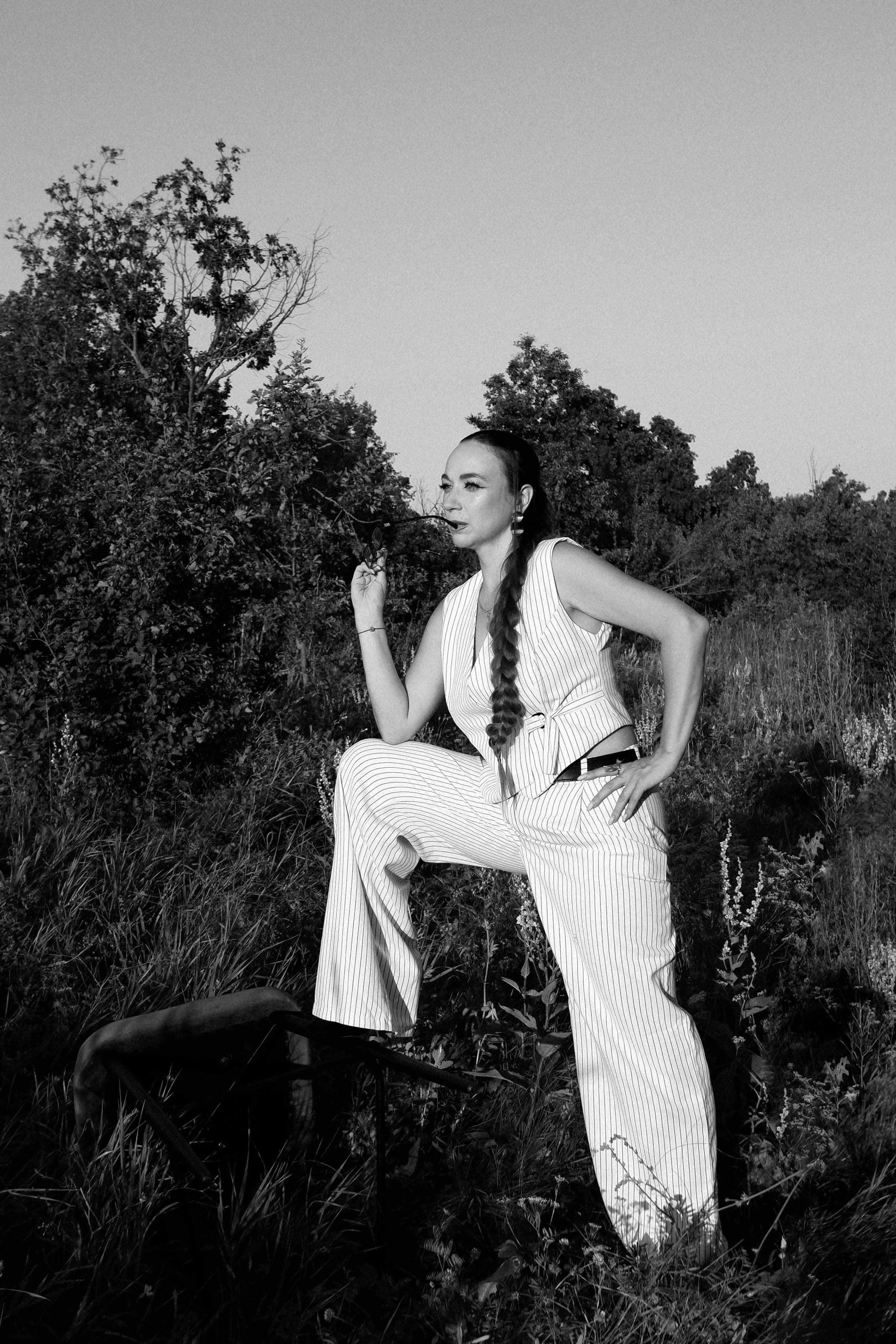 A woman with long braided hair in a striped jumpsuit sitting outdoors on a grassy field, holding glasses near her mouth, with trees and open sky in the background, captured in black and white.