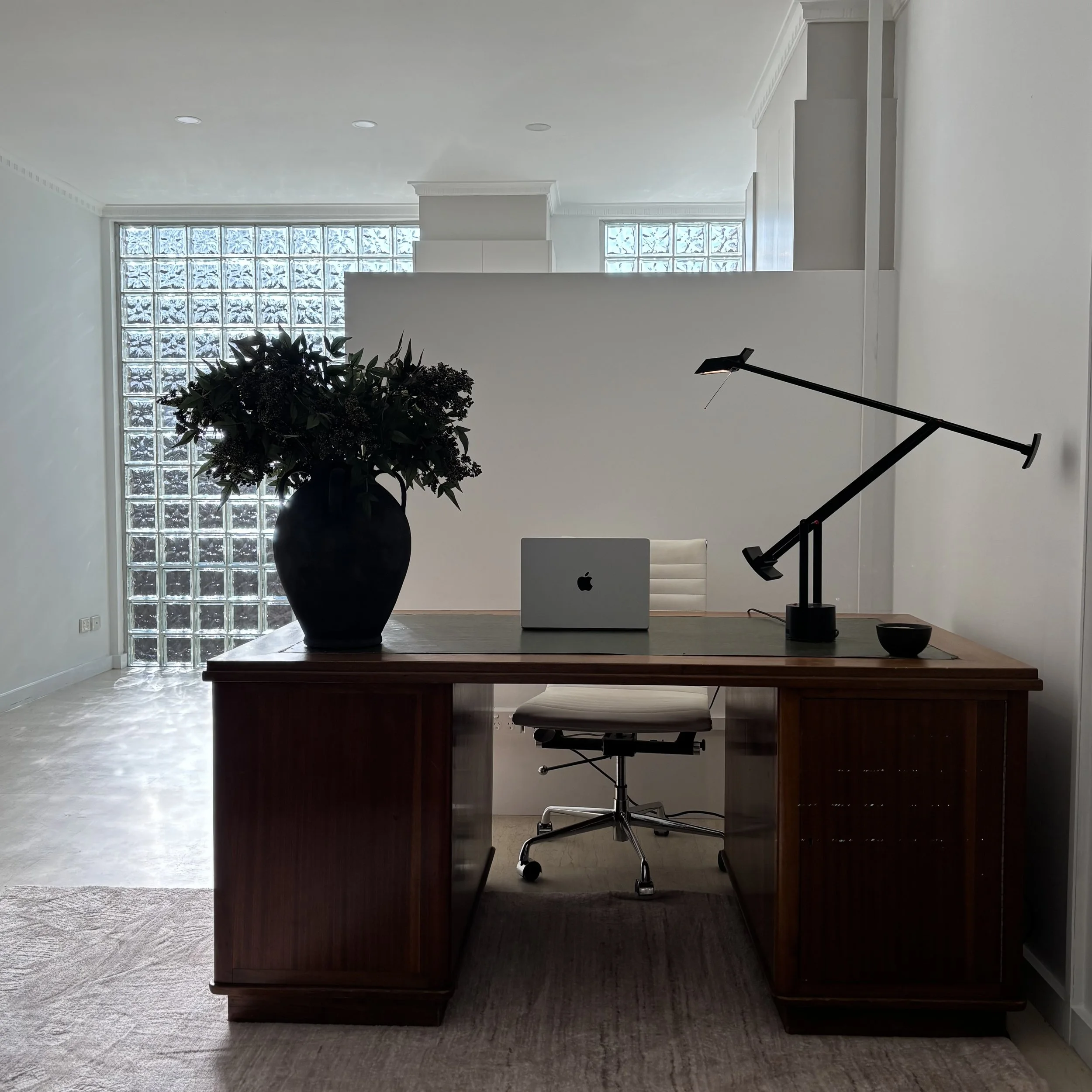 An office space with a wooden desk, a black desk lamp, a chair, a potted plant, a closed laptop, and a white chair, with glass block windows letting in natural light.