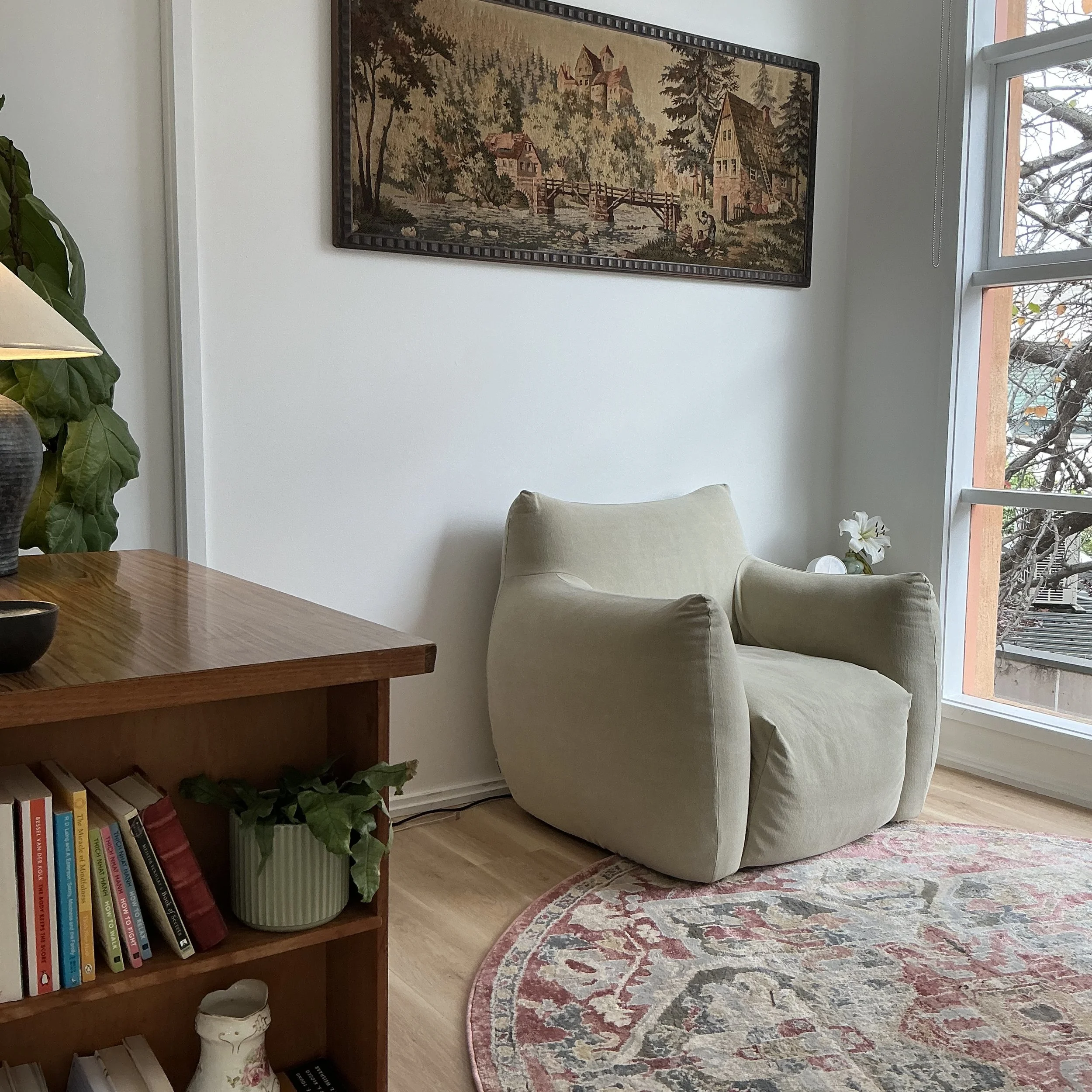 A cozy living room corner with a beige armchair, a wooden bookshelf filled with colorful books, a decorative vase, a large patterned rug, a framed tapestry of a woodland scene on the wall, a window with blinds showing tree branches outside, and a potted plant.