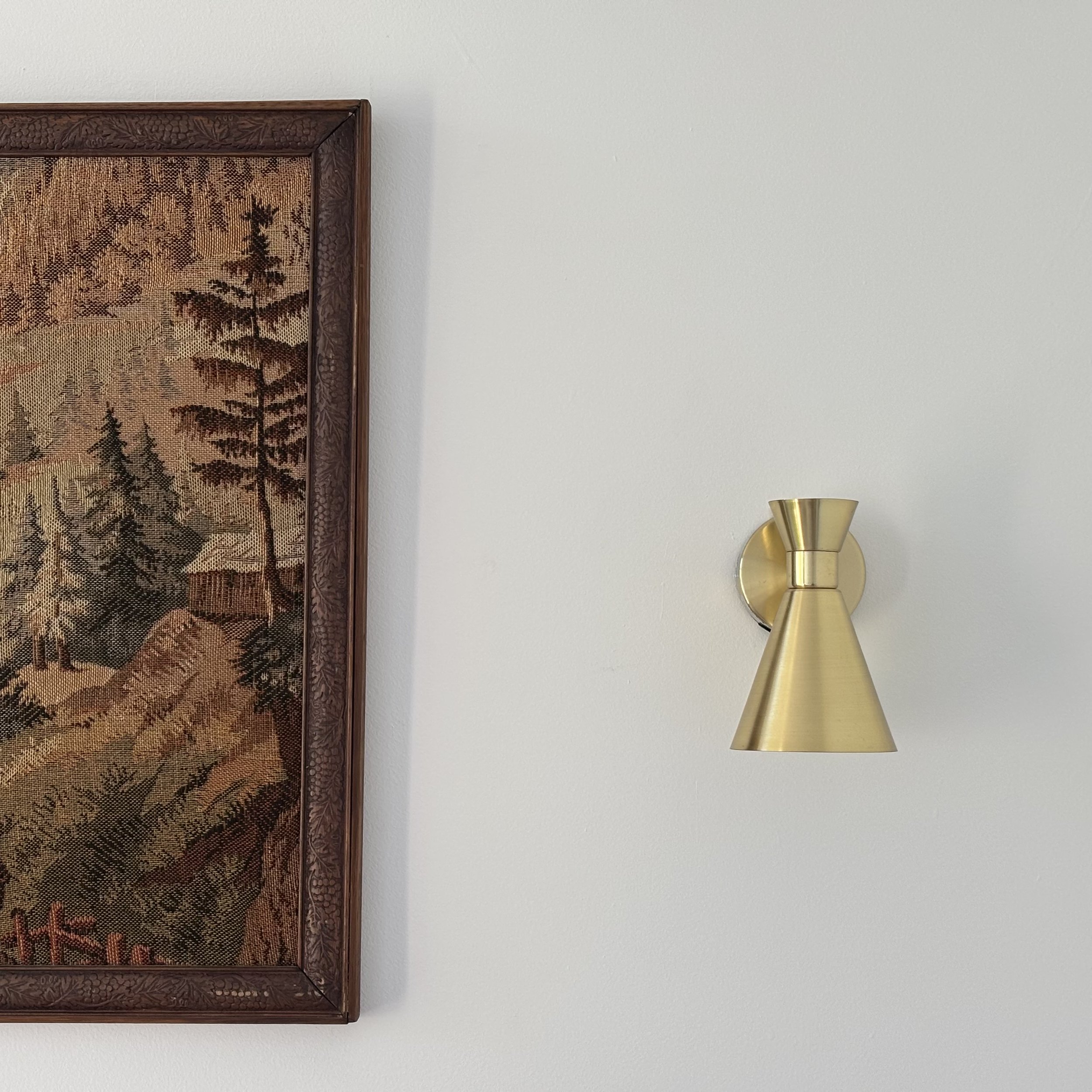 Part of a decorative framed textile artwork with a nature scene, next to a gold-colored wall sconce against a plain white wall.