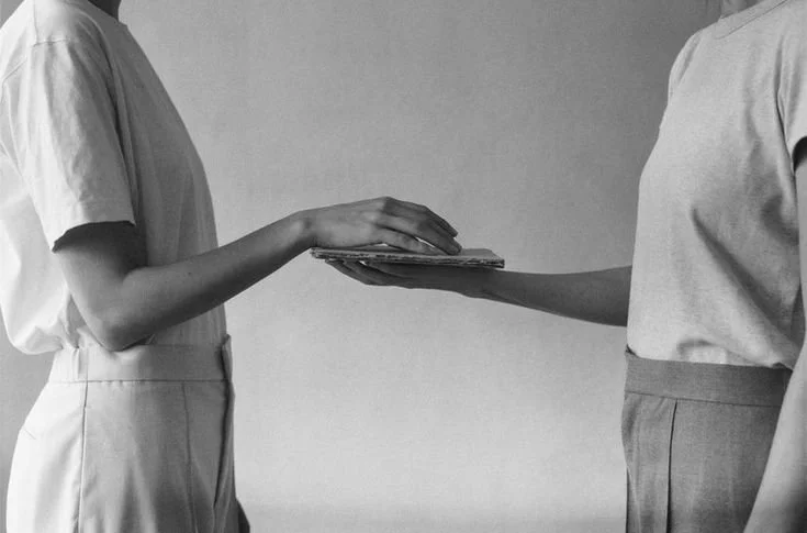 Two individuals exchanging a flat object, such as a folder or book, against a plain wall background.