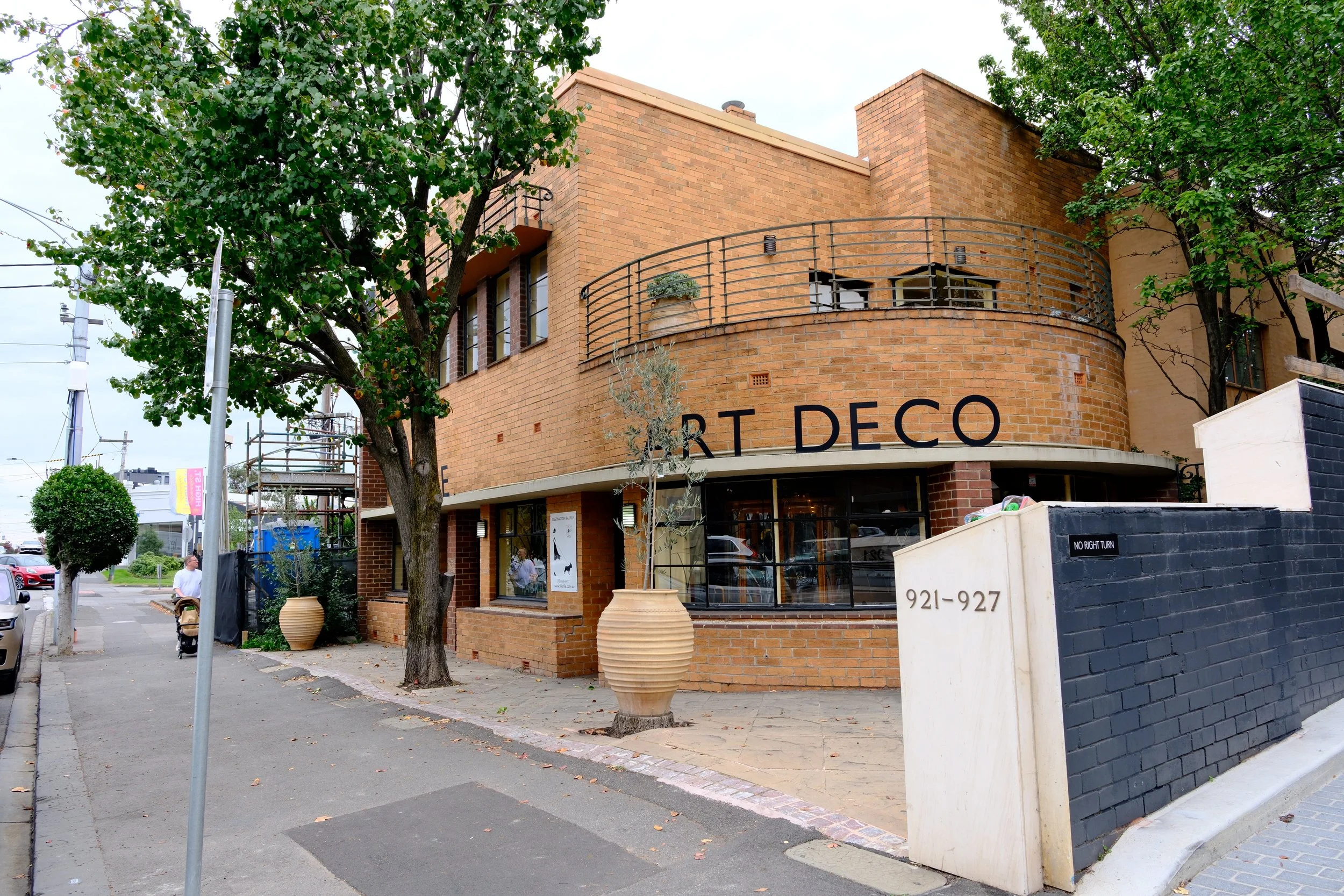 A corner view of a brick building with large windows and an outdoor balcony. There are trees with green leaves partially covering the building, and store signage with the words "ART DECO" is visible on the front. Sidewalk and street with parked cars are in front of the building.