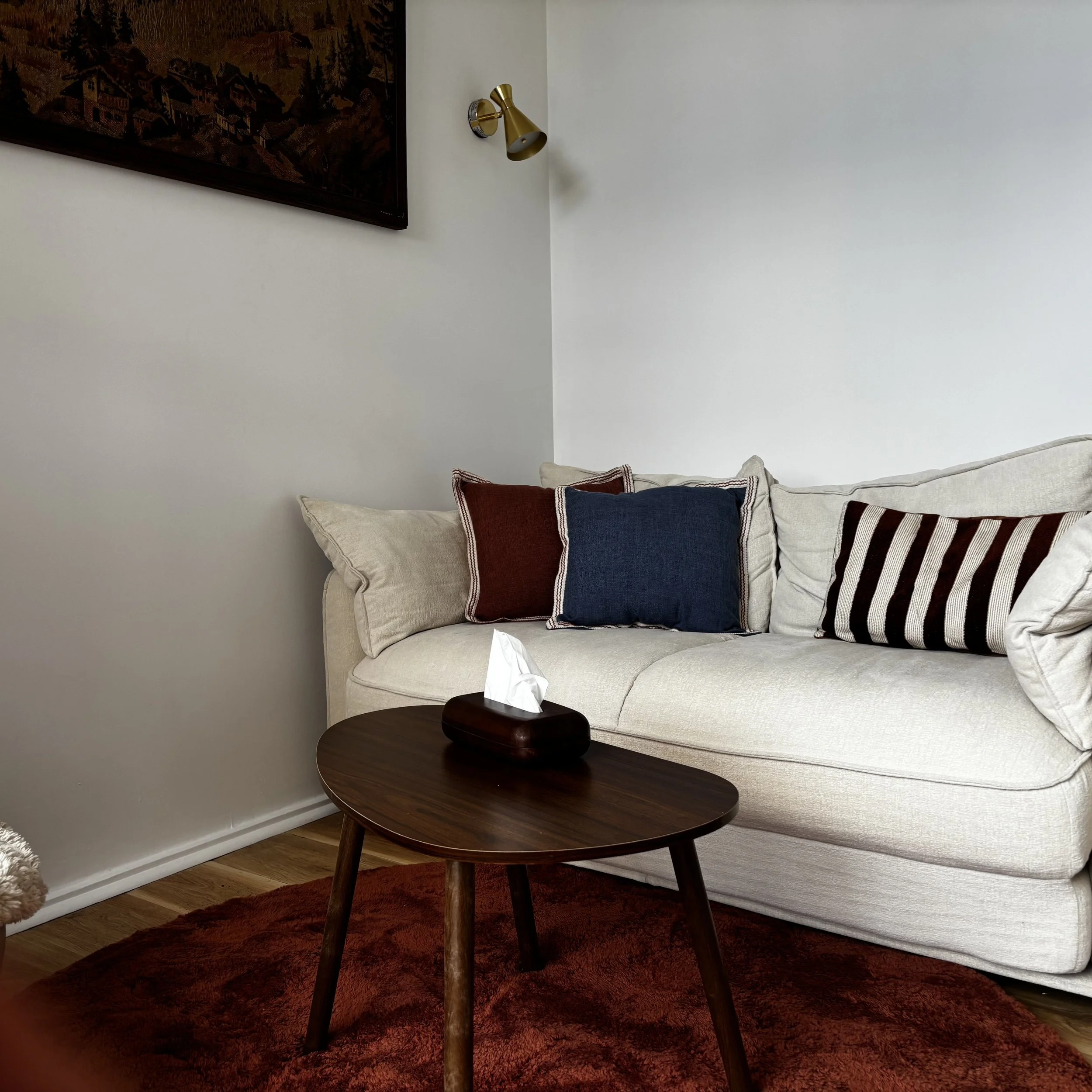 Living room with off-white sofa, wooden table, red rug, pillows, wall art, and wall-mounted lamp.