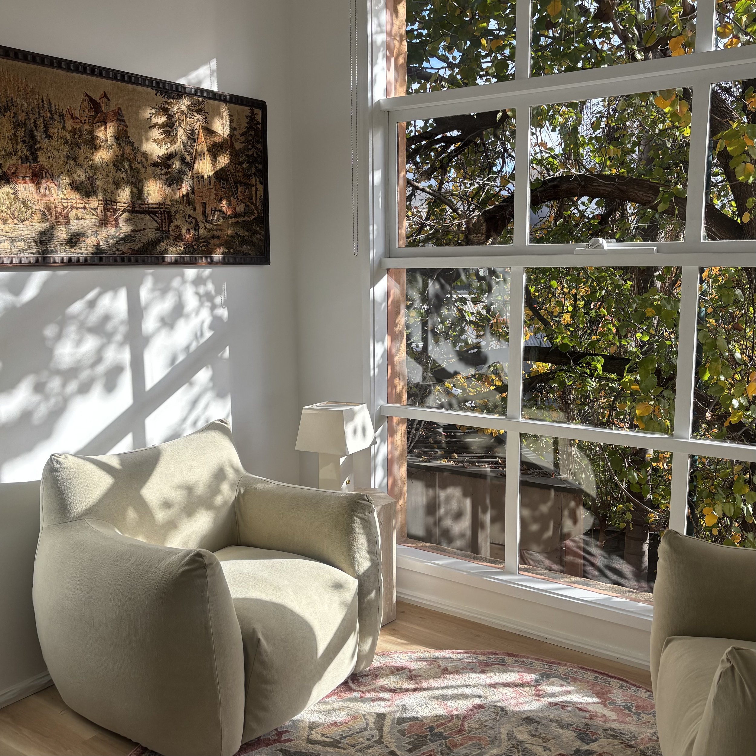 Living room corner with a cream-colored armchair, a small white side table, a colorful area rug, a decorative tapestry on the wall, and large window with white framing revealing a tree with green leaves outside.
