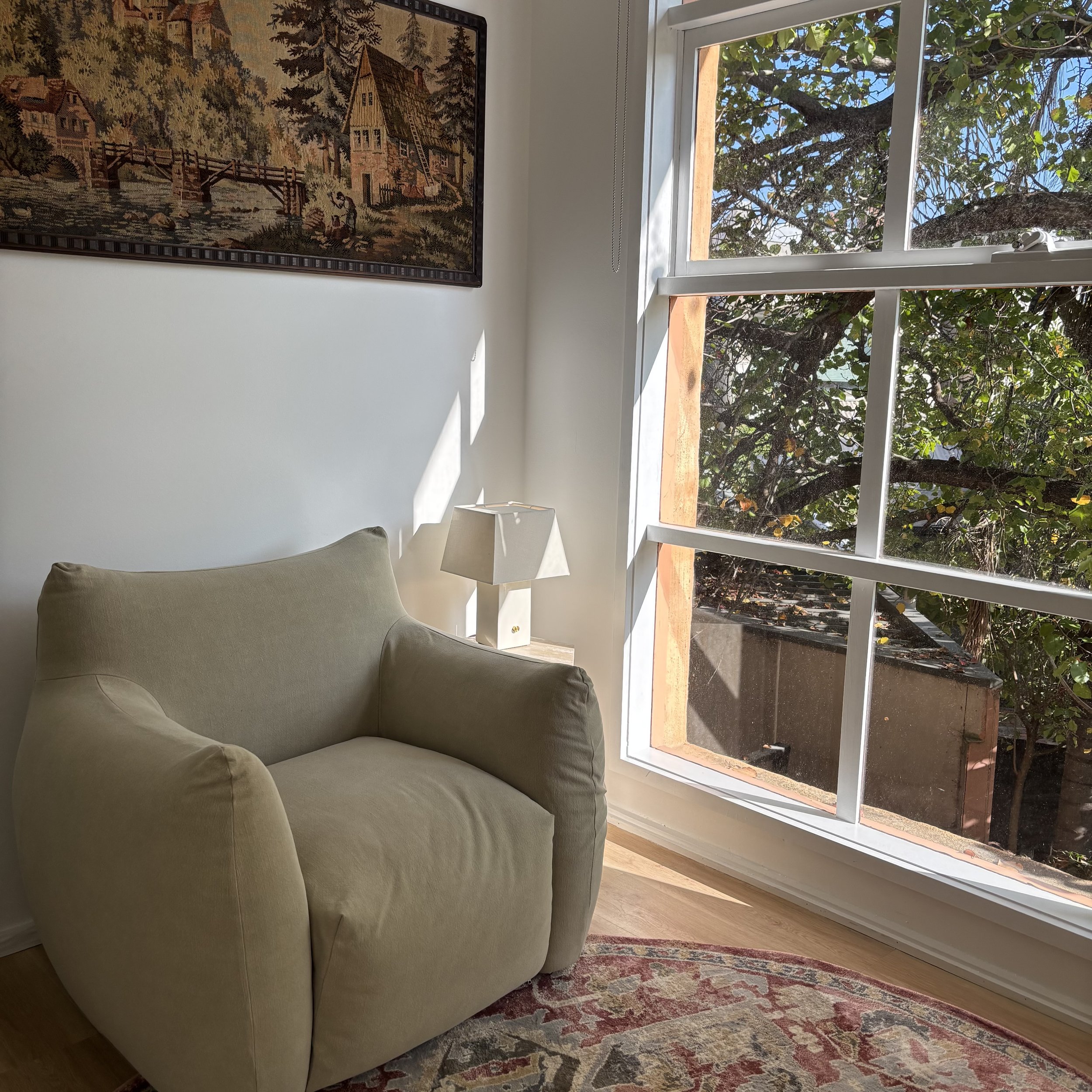 A living room with a large window showing trees outside, a beige sofa, a white lamp on a side table, a framed artwork of a scenic landscape on the wall, and a colorful patterned rug on a wooden floor.