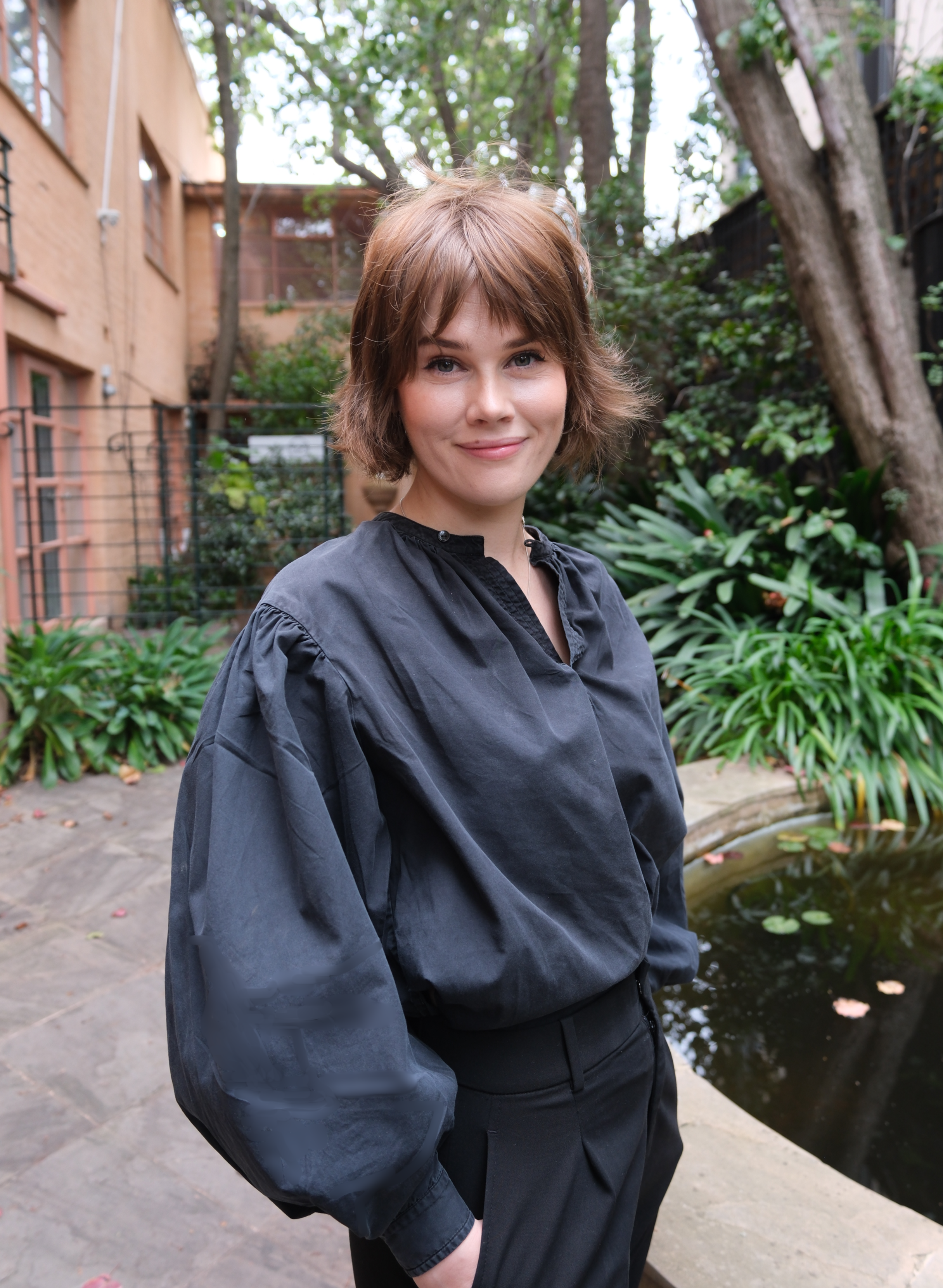 A woman with short brown hair wearing a black jacket and black pants stands outdoors near a pond and lush greenery, smiling at the camera.
