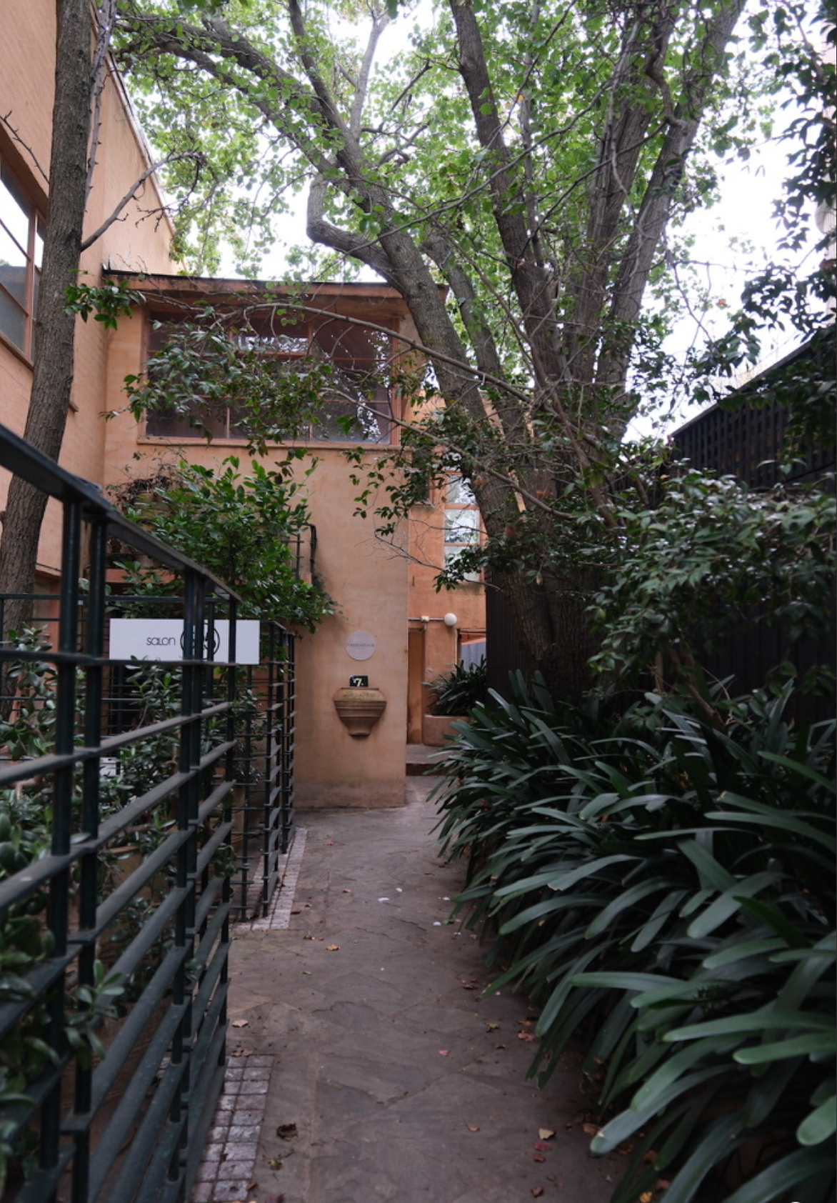 A narrow alleyway surrounded by lush green plants and trees, with a peach-colored building on the left and a black fence on the right. There is a sign on the wall with the word 'salon' and a house number 7.