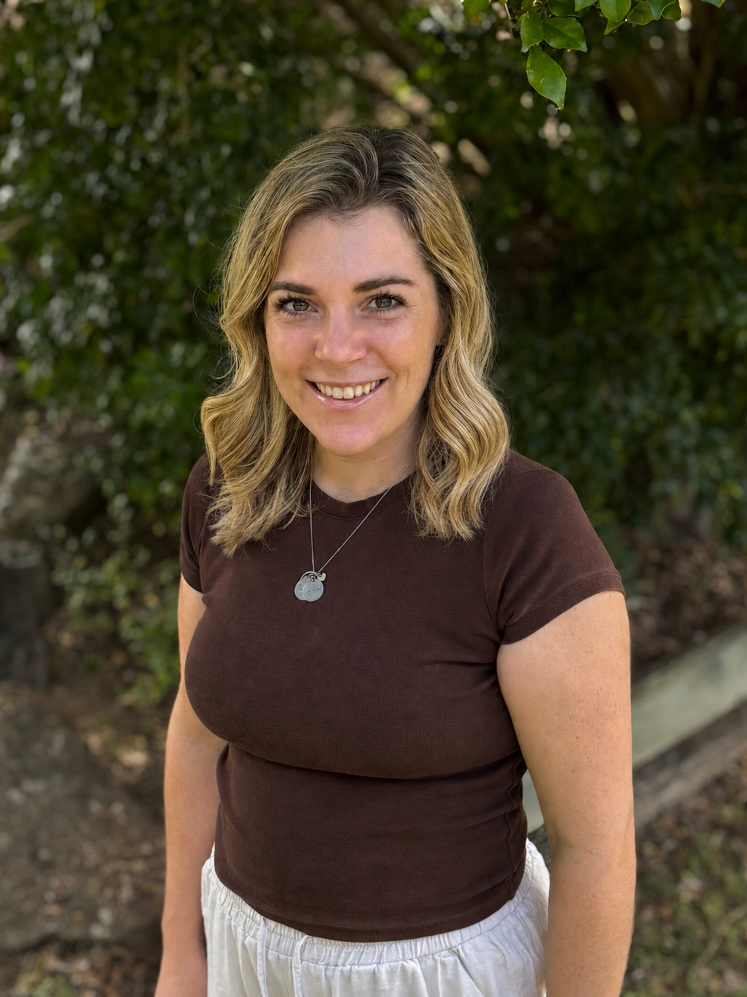 A smiling woman with wavy blonde hair wearing a brown t-shirt and white shorts, standing outdoors with green foliage in the background.
