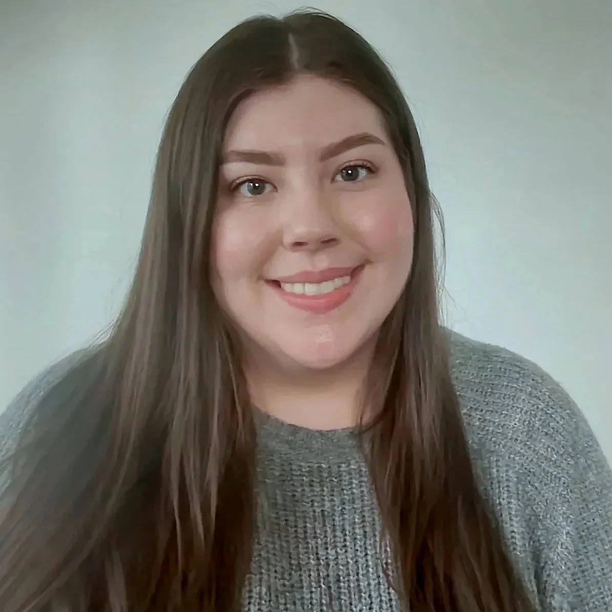 A young woman with long brown hair and light skin, smiling at the camera, wearing a gray knitted sweater against a plain background.