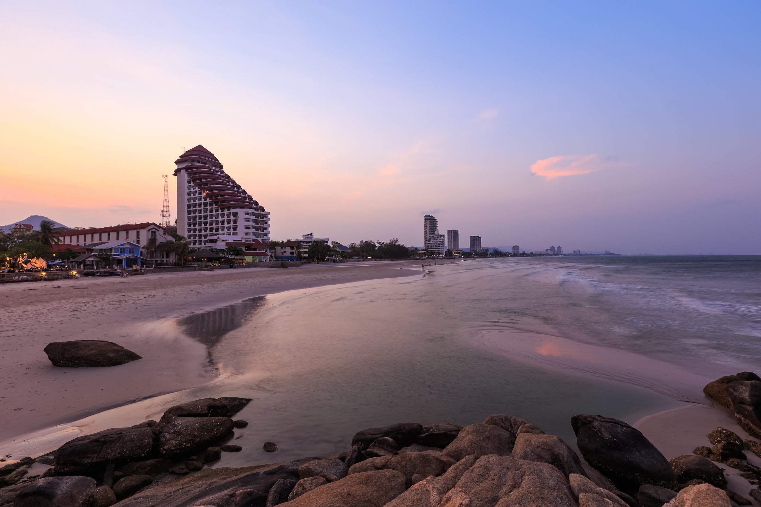 A serene, wide-angle view of a clean Hua Hin beach at sunset, featuring coastal high-rise condominiums and large coastal rocks, symbolizing the safe and peaceful Royal City Lifestyle near premier properties.