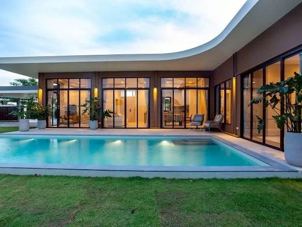 A sophisticated L-shaped modern pool villa featuring expansive glass facades and a pristine turquoise swimming pool set against a backdrop of lush tropical greenery and a clear blue sky.